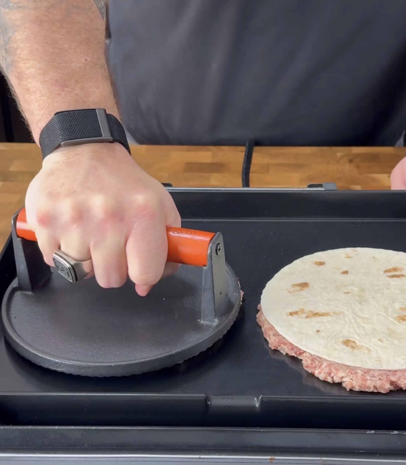 A person presses a burger patty with a metal press on a griddle while another patty topped with a tortilla cooks nearby. The person wears a black ring and a smartwatch.