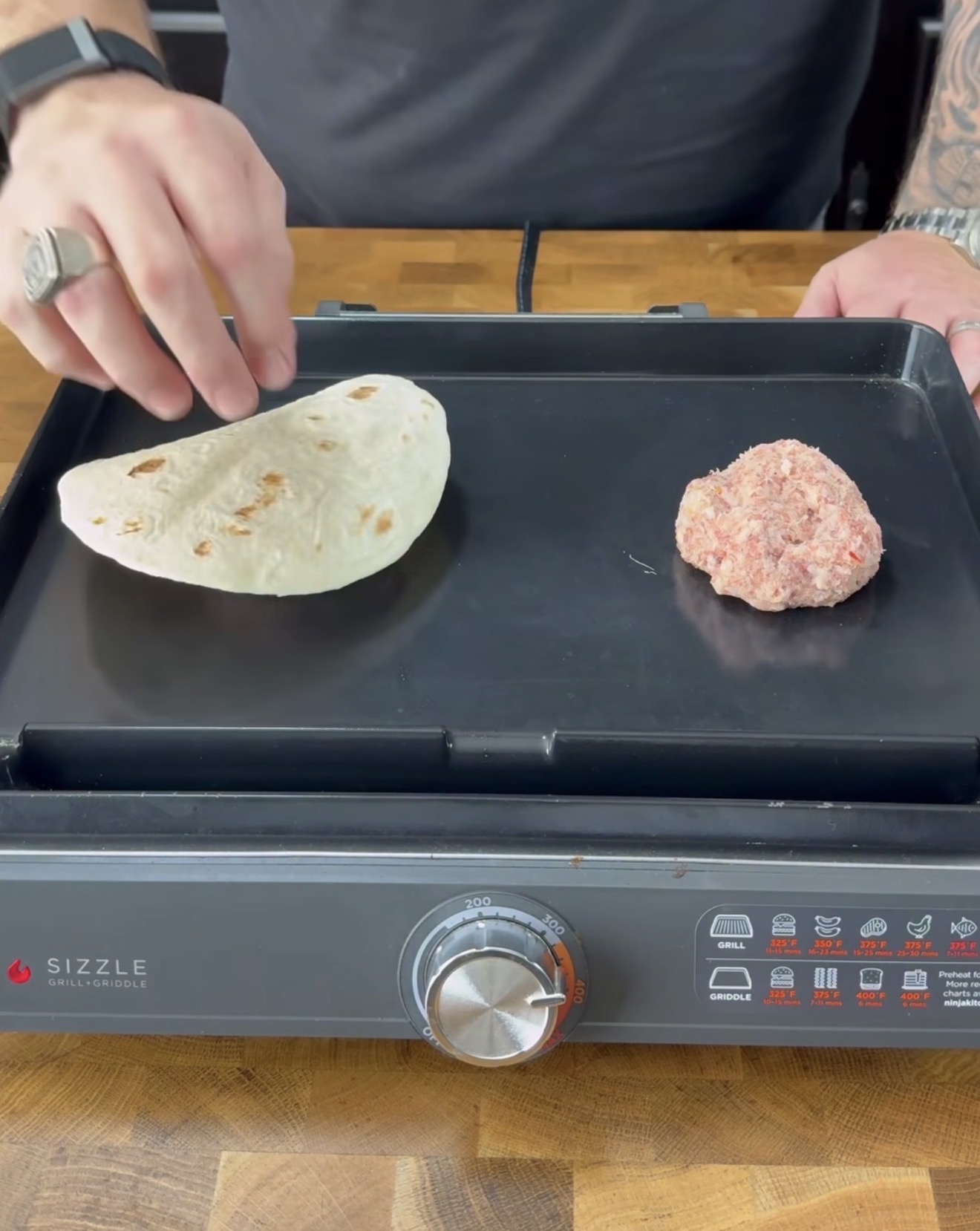 A person places a folded tortilla and a ball of raw ground meat on a Sizzle griddle, preparing to cook. Their hands and arms, which have tattoos and a ring, are visible. The griddle is on a wooden countertop.