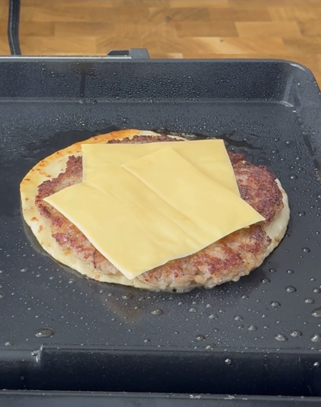 A large round patty topped with two slices of yellow cheese is cooking on a black griddle, with oil and steam visible, and a wooden surface in the background.