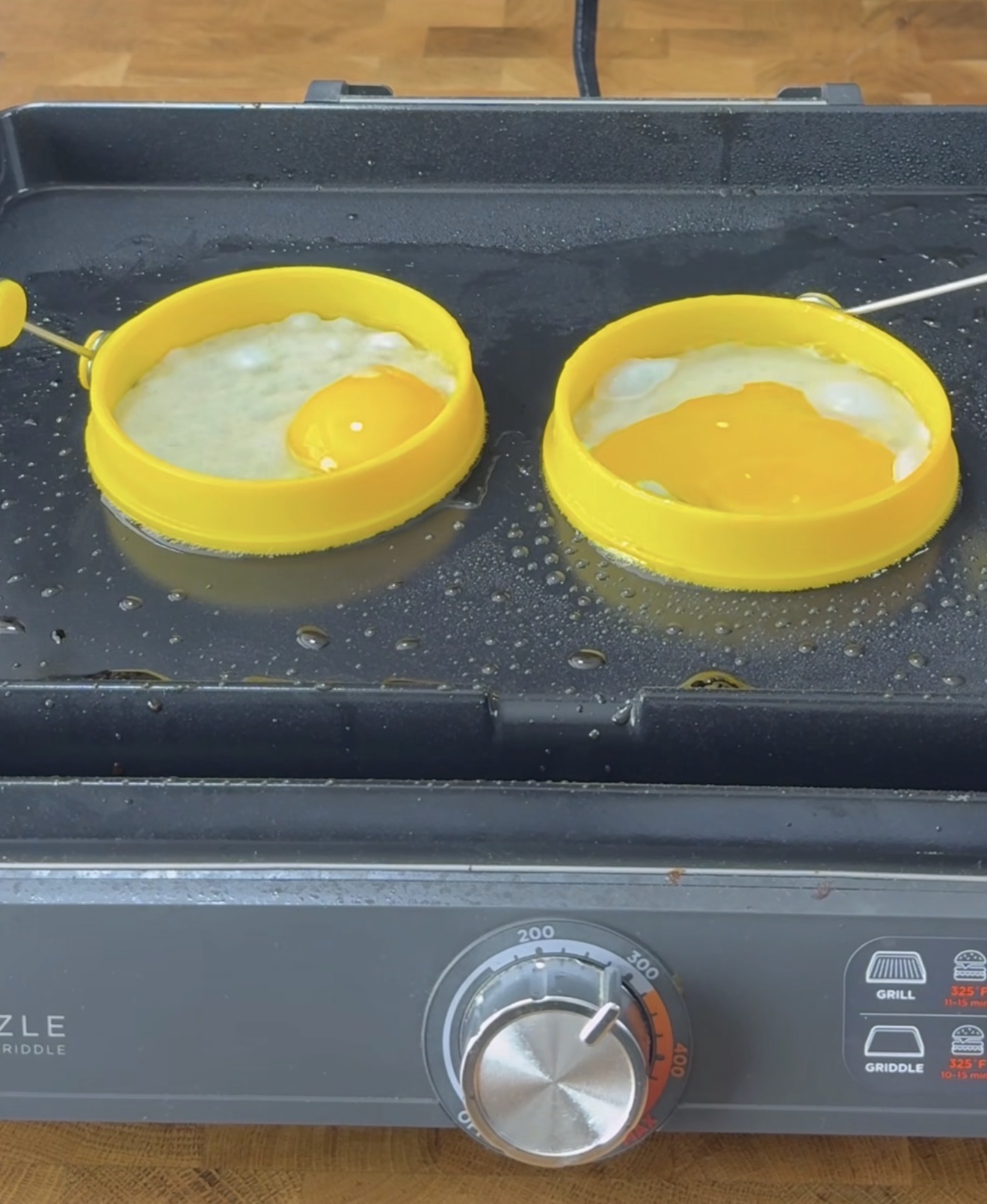 Two eggs are being cooked on a griddle inside yellow silicone egg rings; one yolk is broken, while the other is intact. The surface is oily, and the griddle dial is set to about 200 degrees.