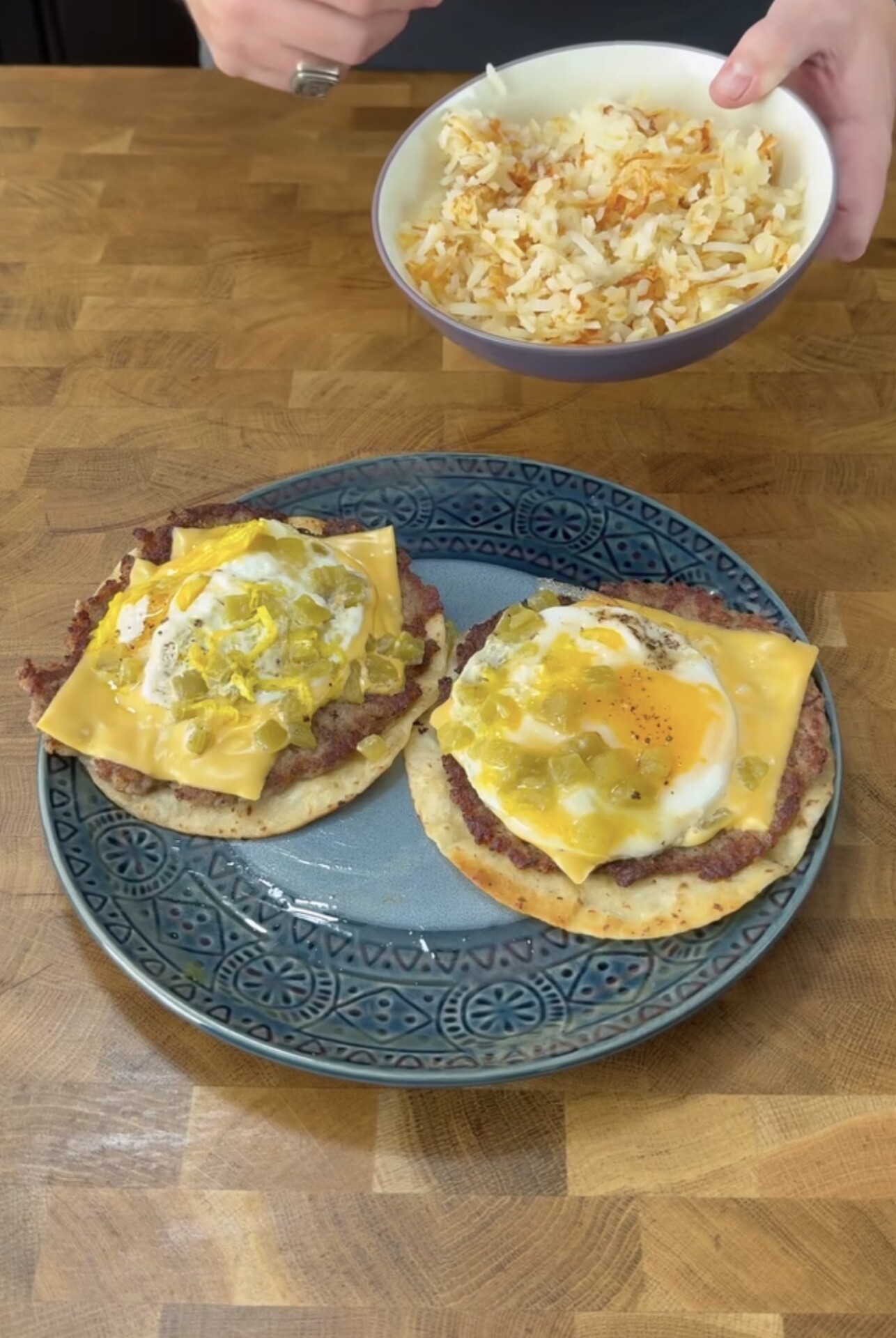 A blue plate holds two flatbreads topped with refried beans, cheddar cheese, eggs, and green chiles. In the background, a hand holds a bowl of rice over a wooden surface.