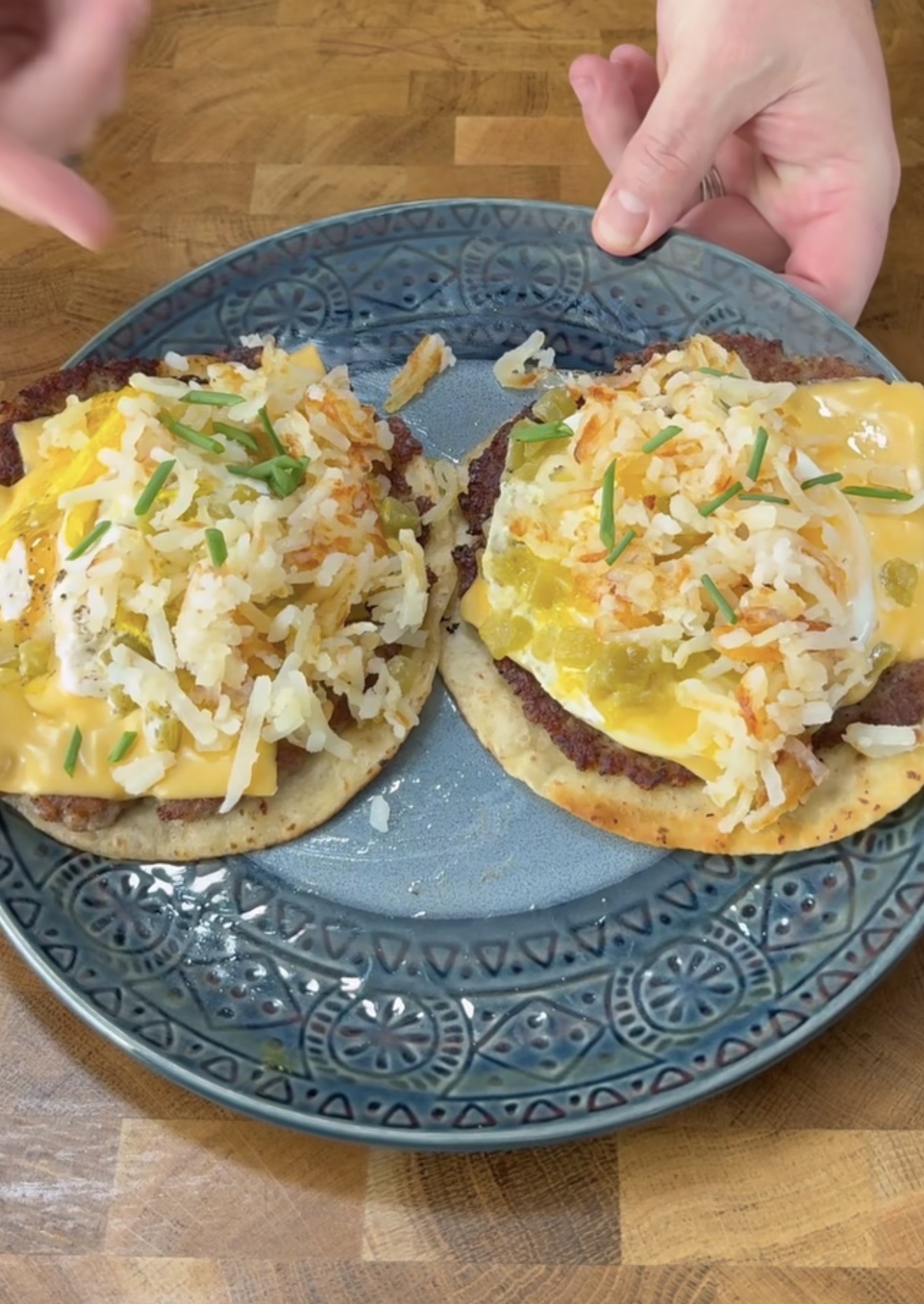 A blue decorative plate holds two open-faced breakfast sandwiches with sausage patties, melted cheese, fried eggs, hash browns, and chopped chives. Two hands are positioned near the plate.