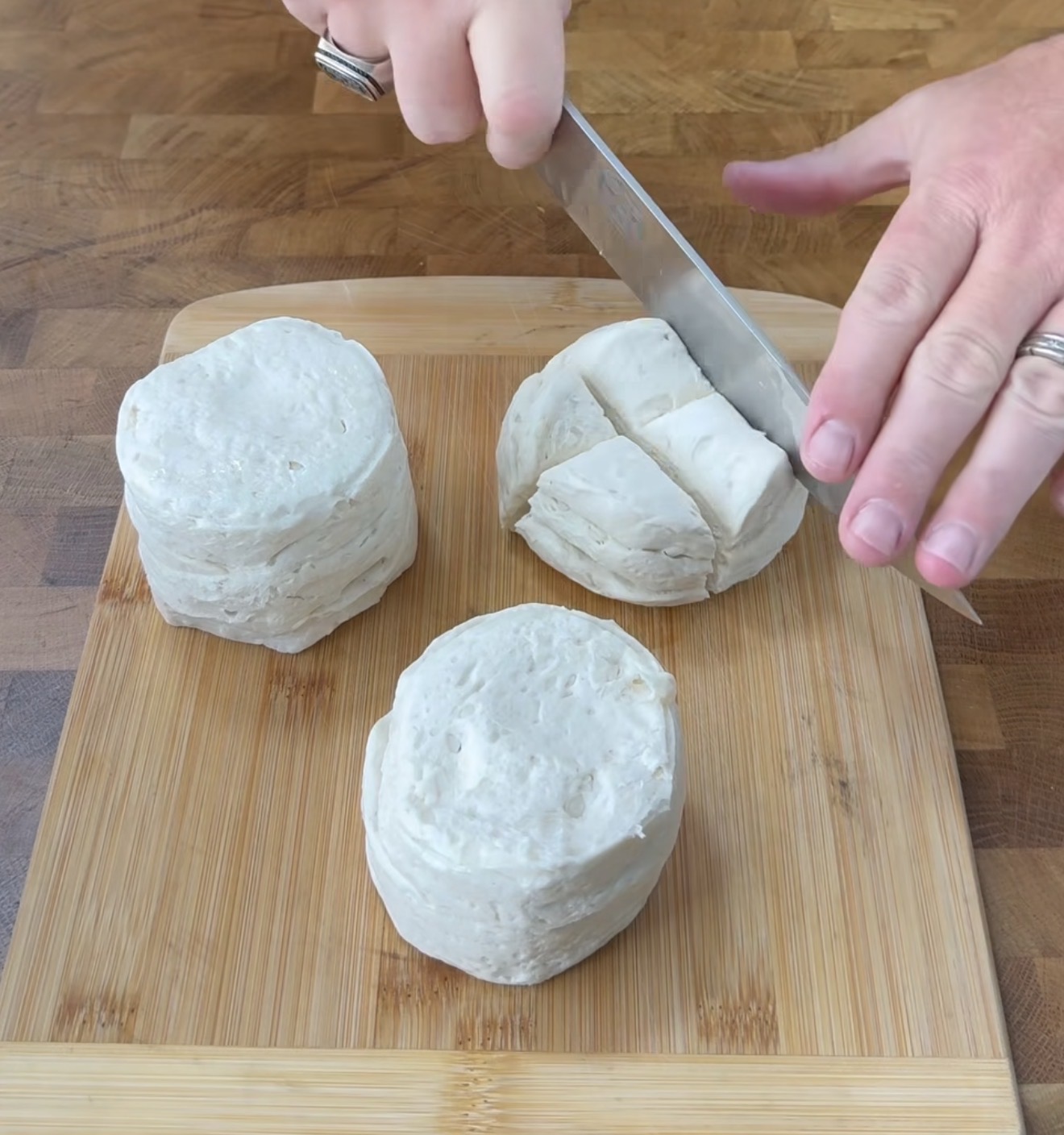 A person’s hands are slicing biscuit dough on a wooden cutting board, dividing one round piece into smaller sections. Two more whole pieces of biscuit dough are next to the one being cut.