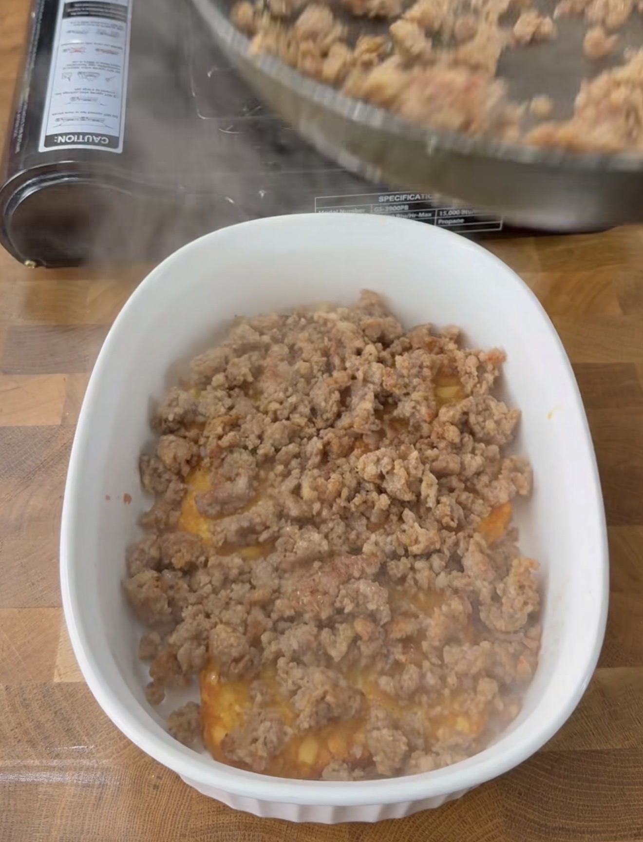Cooked ground meat being transferred from a pan into a white casserole dish on a wooden countertop, with steam rising from the dish.