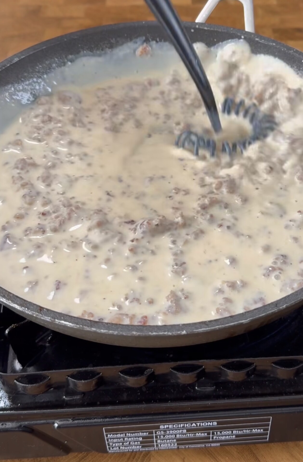 A pan on a portable stove contains ground meat cooking in creamy gravy, being stirred with a black utensil.
