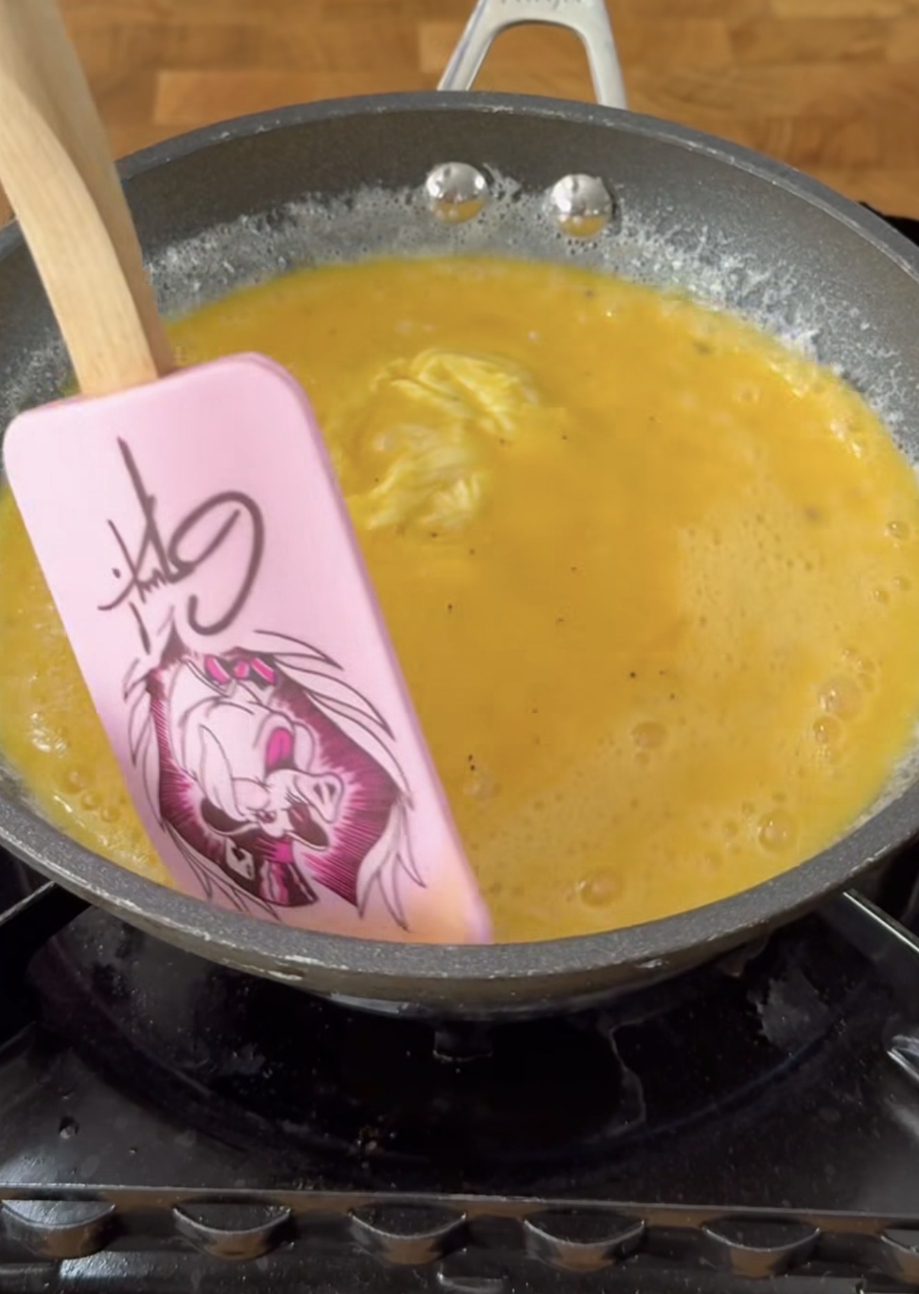 A nonstick pan with beaten eggs cooking on a stove. A pink spatula with an anime-style character illustration is stirring the eggs.