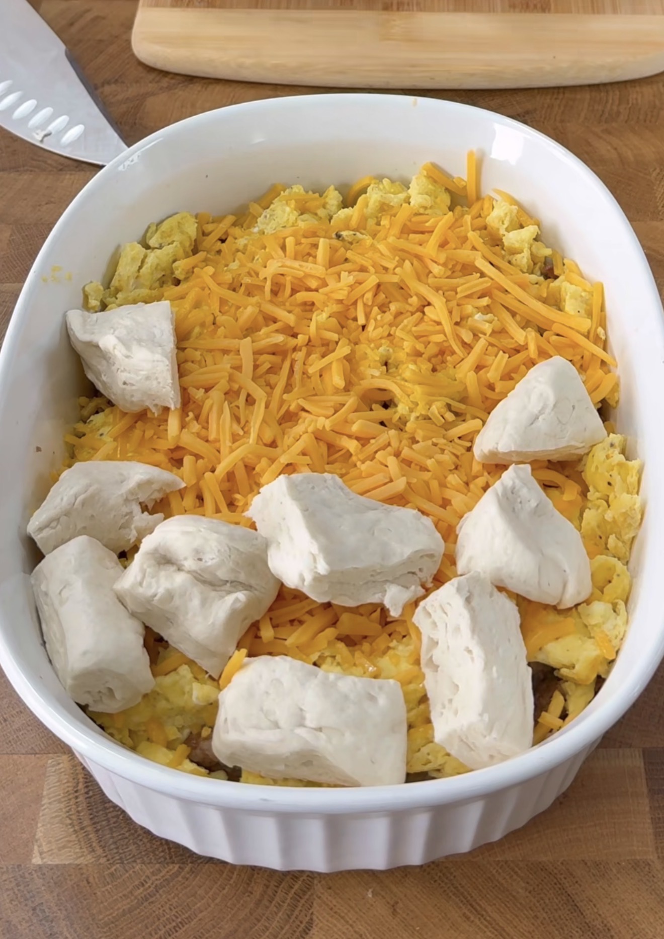 A white baking dish contains a mixture of scrambled eggs topped with shredded cheddar cheese and several uncooked biscuit dough pieces scattered on top. The dish sits on a wooden surface.