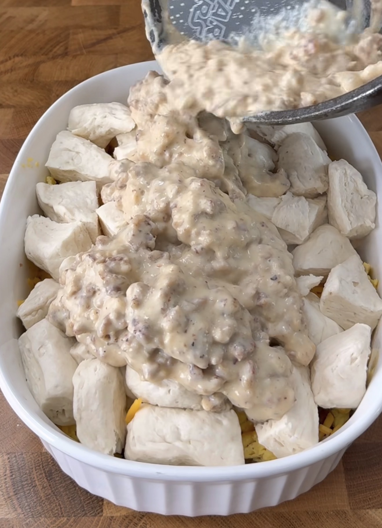 A white baking dish filled with biscuit dough pieces is being topped with a creamy sausage gravy mixture, poured from a pan, on a wooden countertop.