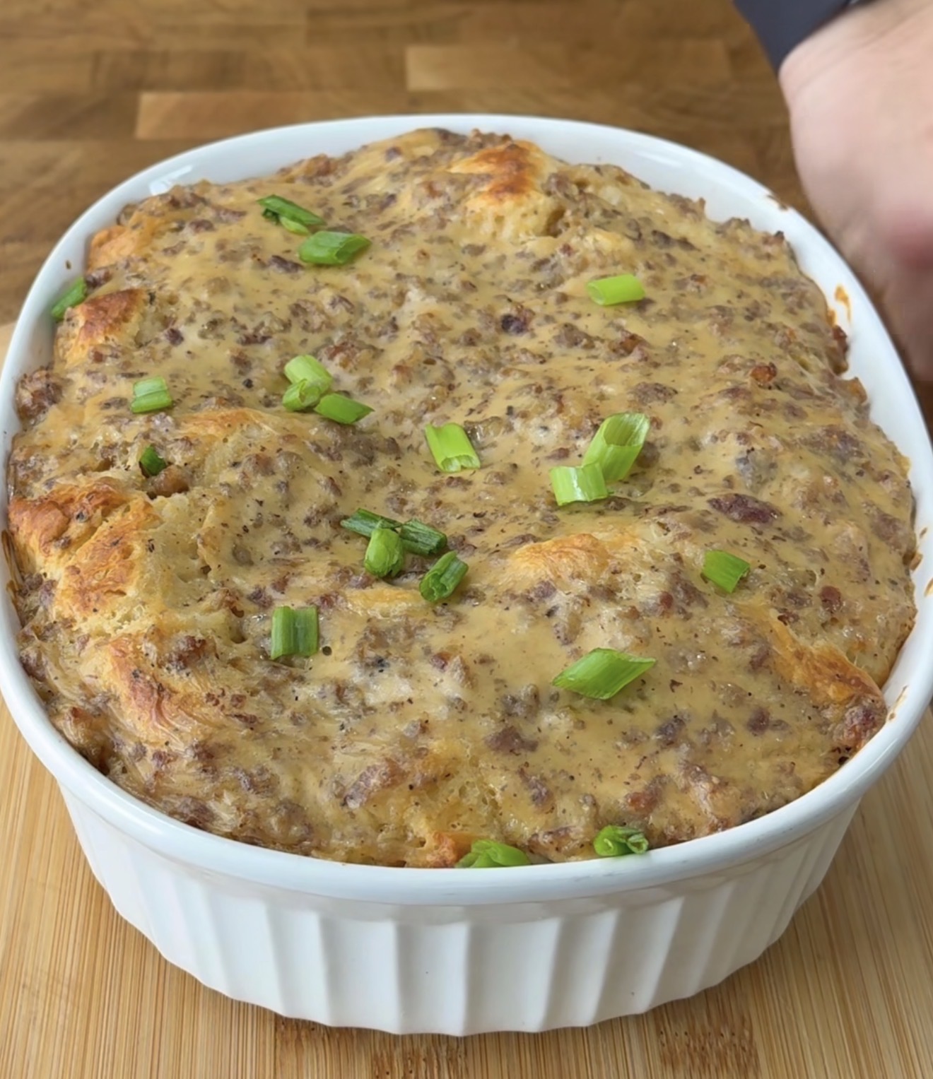 A baked casserole in a white dish, topped with browned, cheesy sausage mixture and garnished with chopped green onions, sits on a wooden surface.