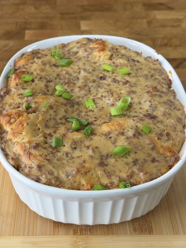 A casserole dish filled with a baked savory dish topped with browned sausage and cheese, garnished with chopped green onions, sits on a wooden surface.