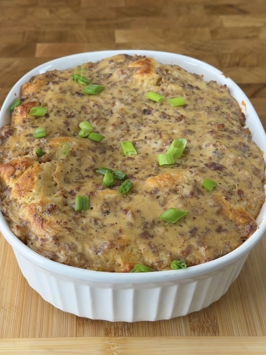 A casserole dish filled with a baked savory dish topped with browned sausage and cheese, garnished with chopped green onions, sits on a wooden surface.