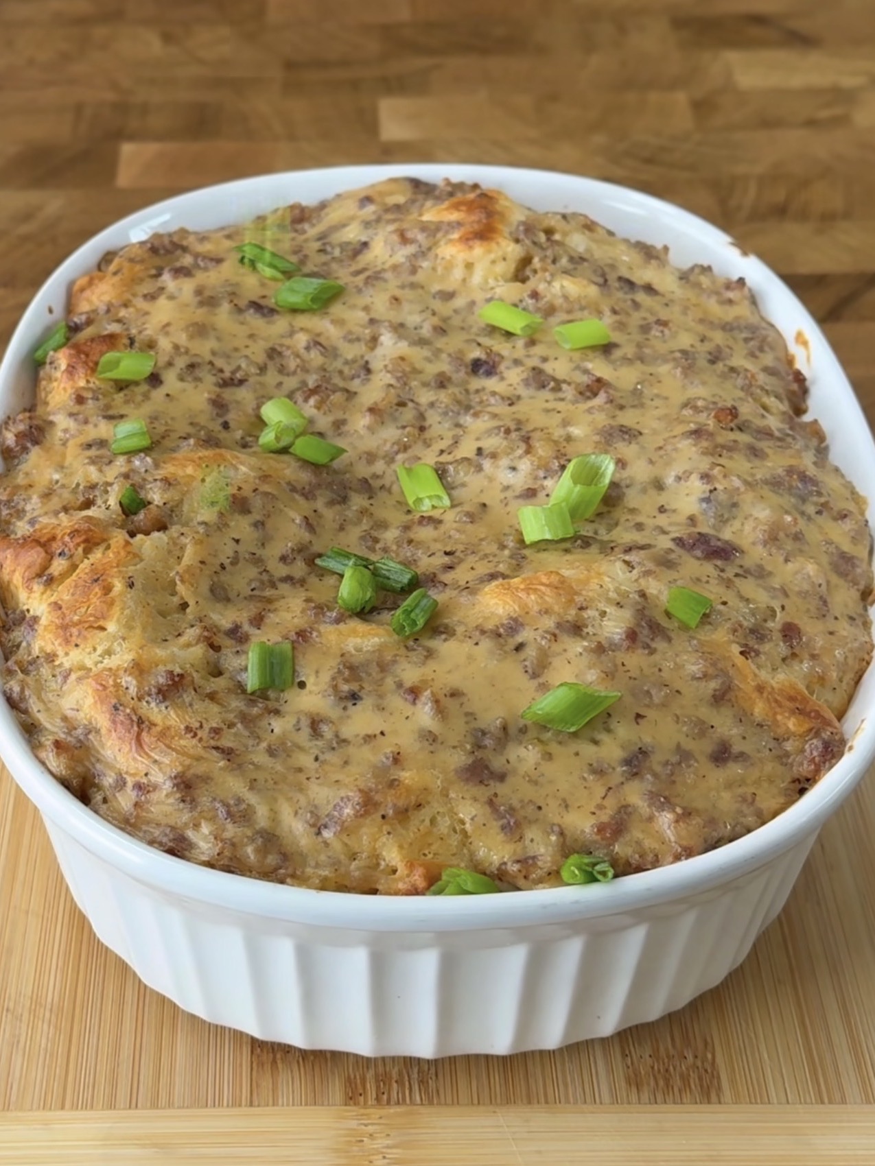 A casserole dish filled with a baked savory dish topped with browned sausage and cheese, garnished with chopped green onions, sits on a wooden surface.