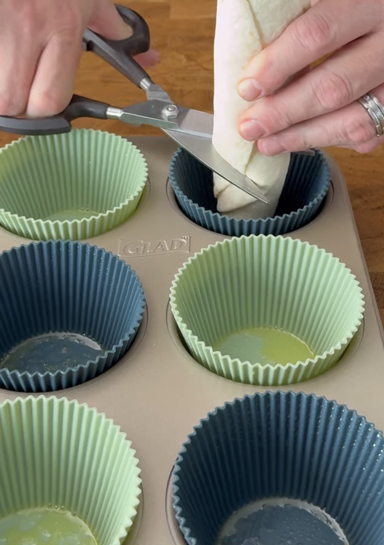 A person uses scissors to cut a rolled tortilla over a muffin tin lined with silicone baking cups. The tin sits on a wooden surface.