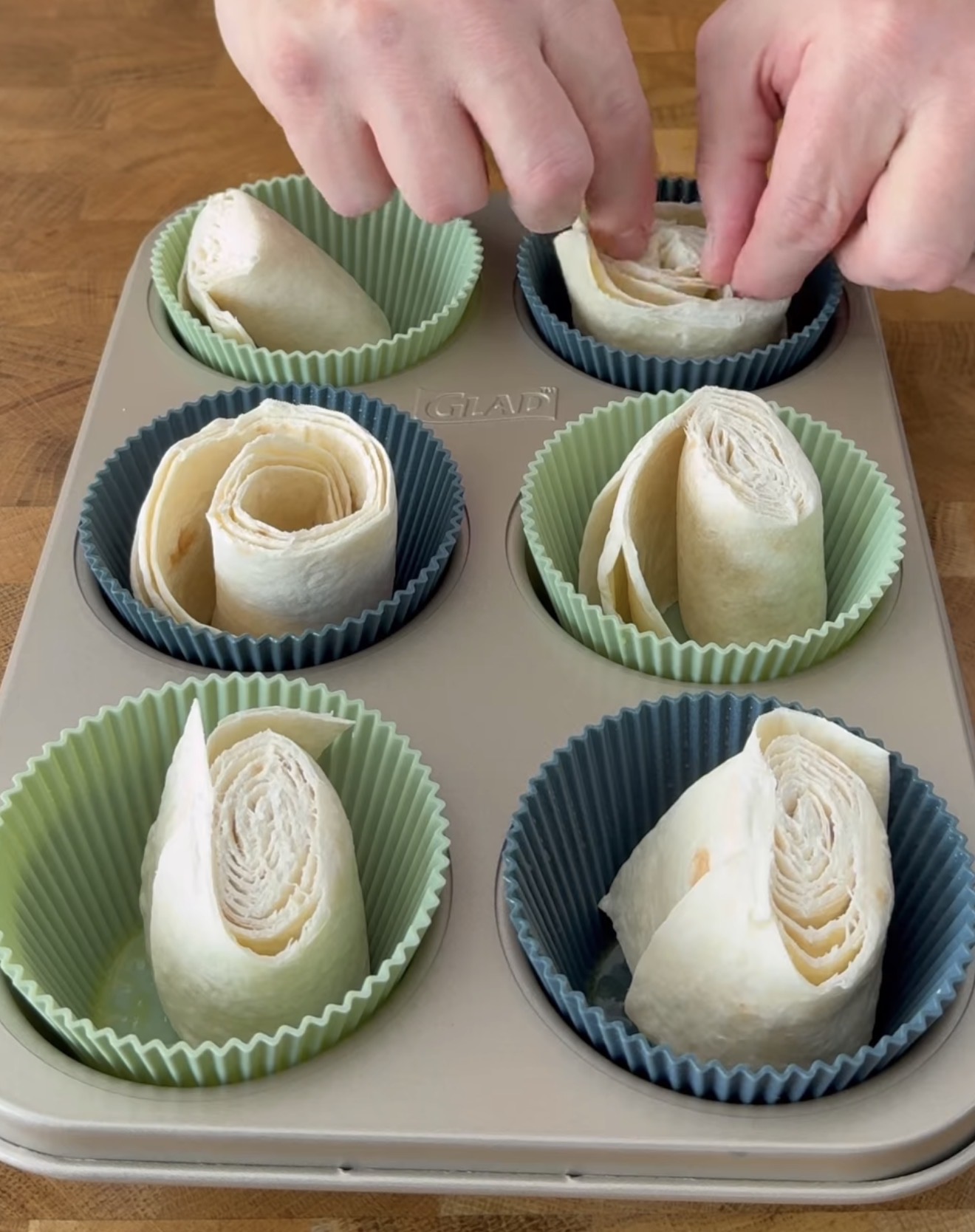 Hands are placing rolled sheets of dough or pastry into silicone muffin cups in a muffin tin. The rolls are arranged upright and the scene is set on a wooden surface.