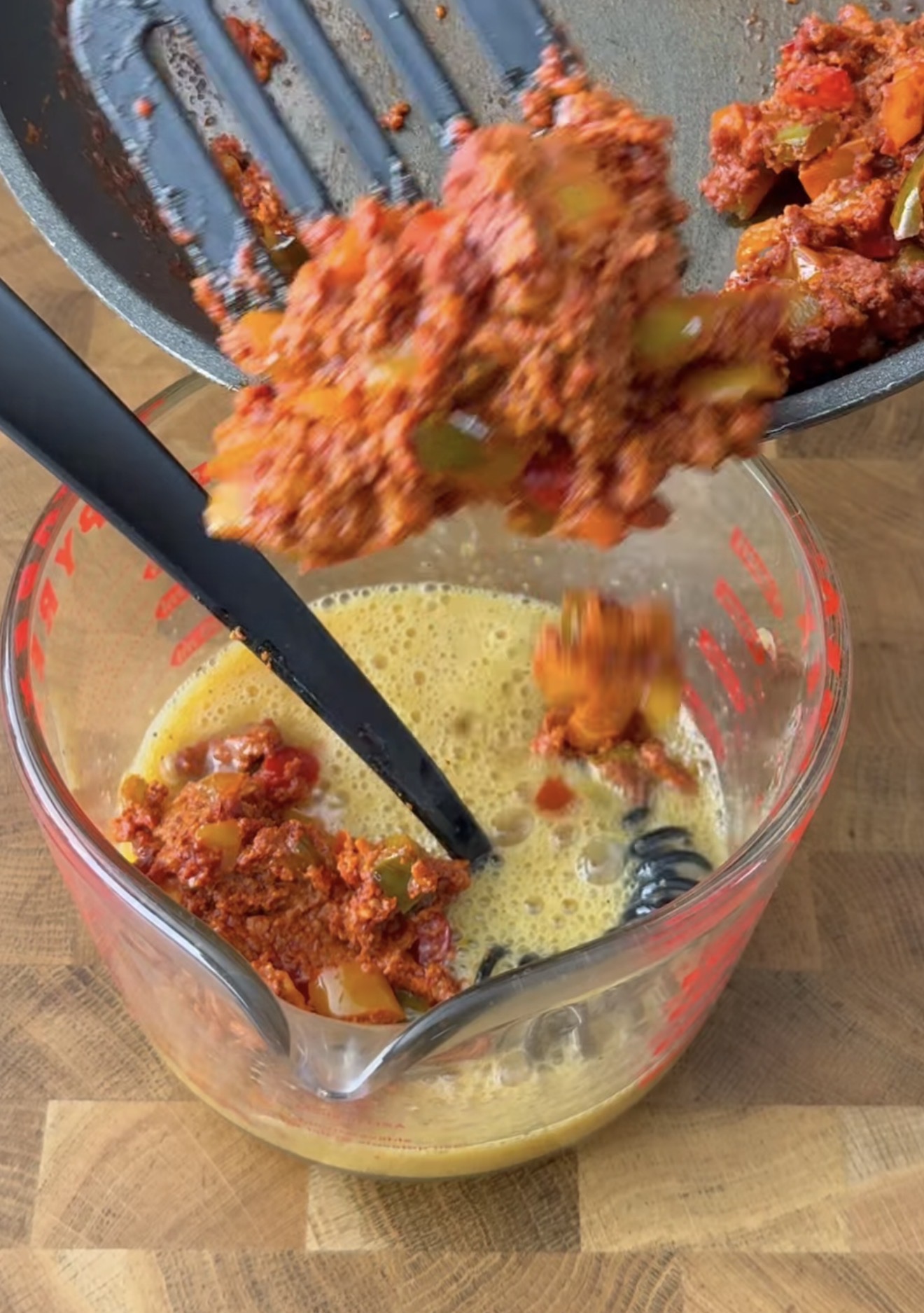 A black spatula transfers cooked chorizo and vegetables from a skillet into a glass measuring cup filled with beaten eggs, all on a wooden countertop.