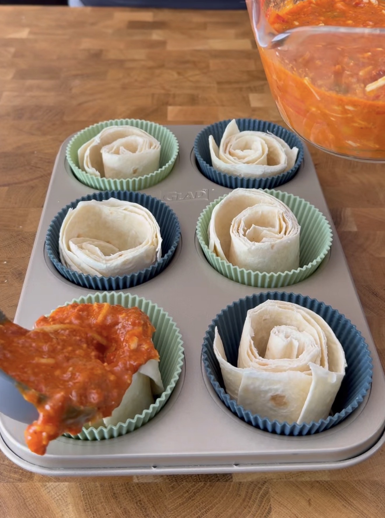 A muffin tray holds six silicone liners, each with rolled tortilla wraps. A spoon is spreading red sauce onto one of the tortillas, with a bowl of sauce visible in the upper right corner.