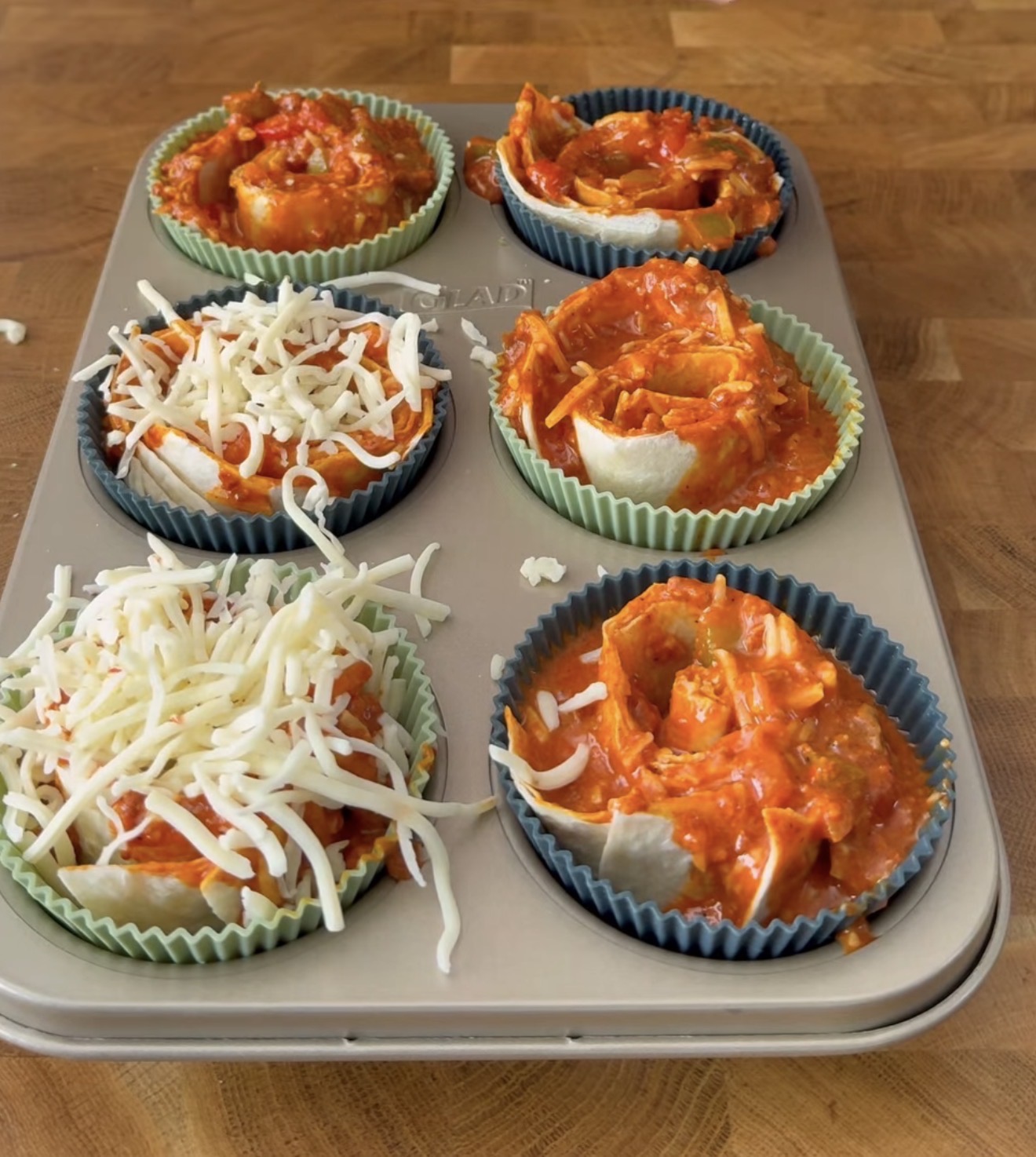 A muffin tin with six colorful silicone liners, each filled with lasagna ingredients like pasta, red sauce, and shredded cheese, ready to be baked on a wooden countertop.