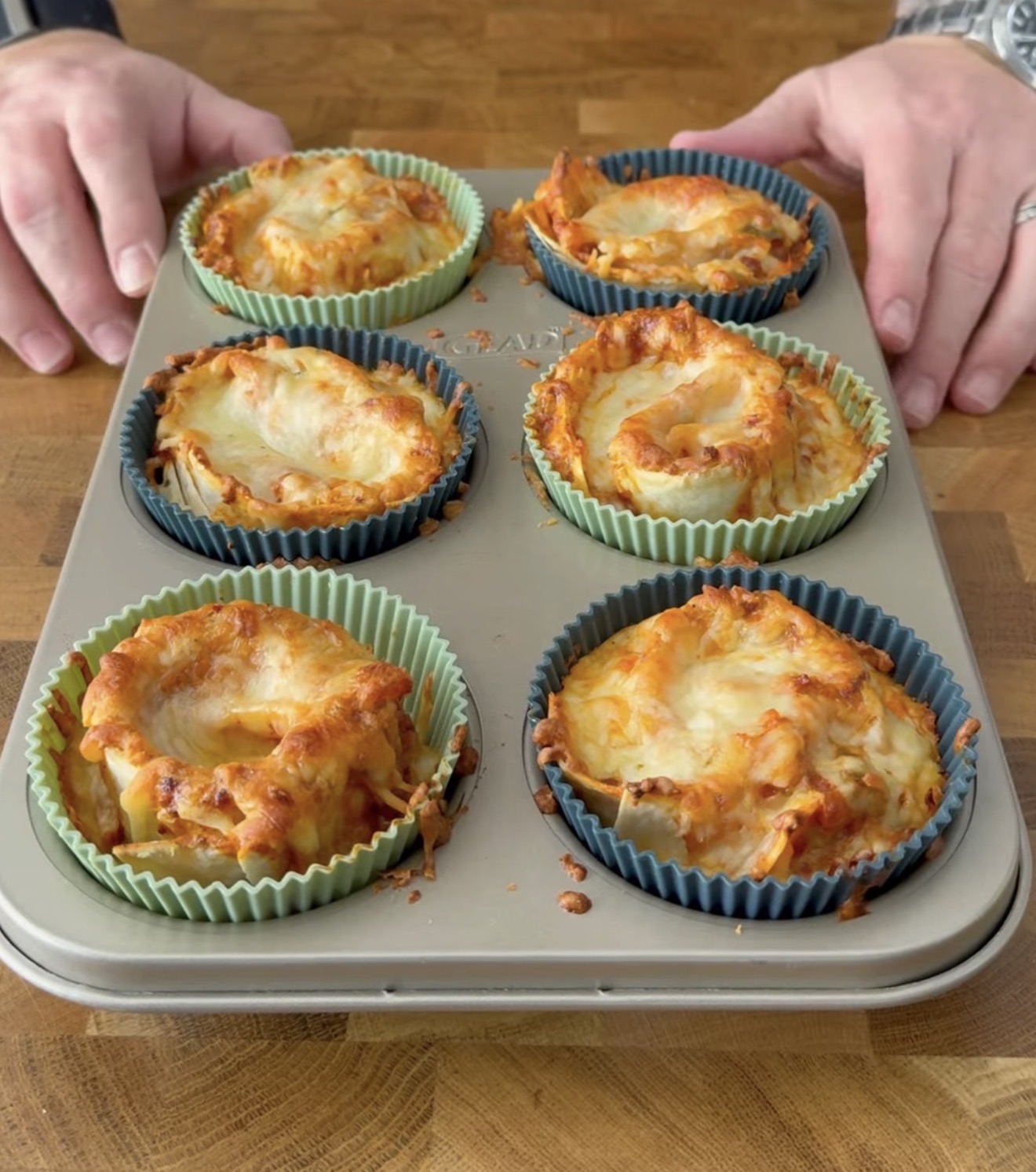 A person holds a muffin pan filled with six cheesy baked pasta cups in colorful silicone liners on a wooden surface.