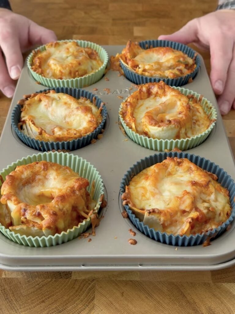 Six baked lasagna cups with melted cheese sit in silicone liners in a muffin pan on a wooden surface, with two hands holding the pan.