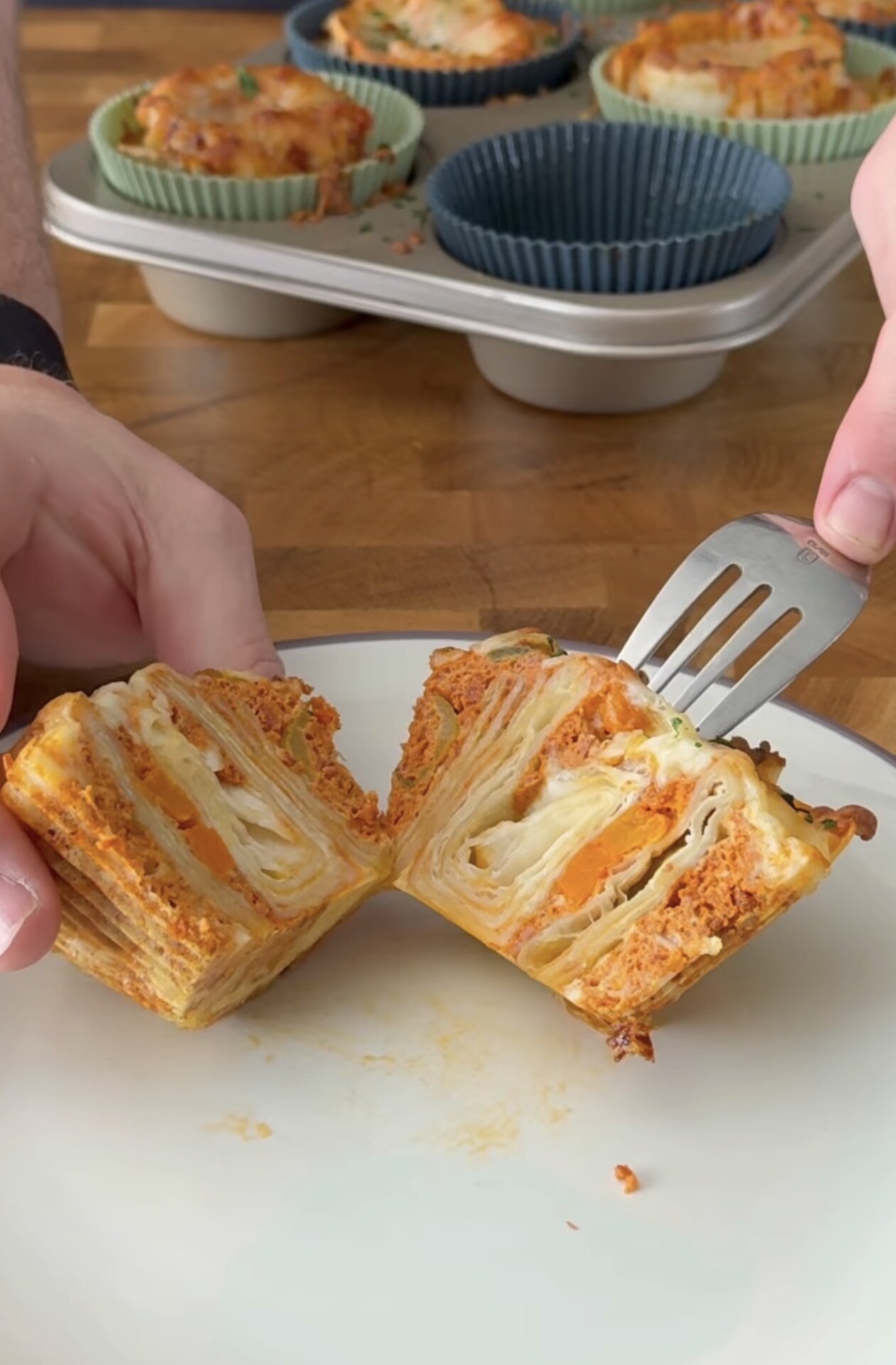 A person uses a fork and hand to split open a single-serve, layered pasta dish with sauce, showing its many visible layers. In the background, similar pasta dishes are in silicone muffin liners in a baking tray.