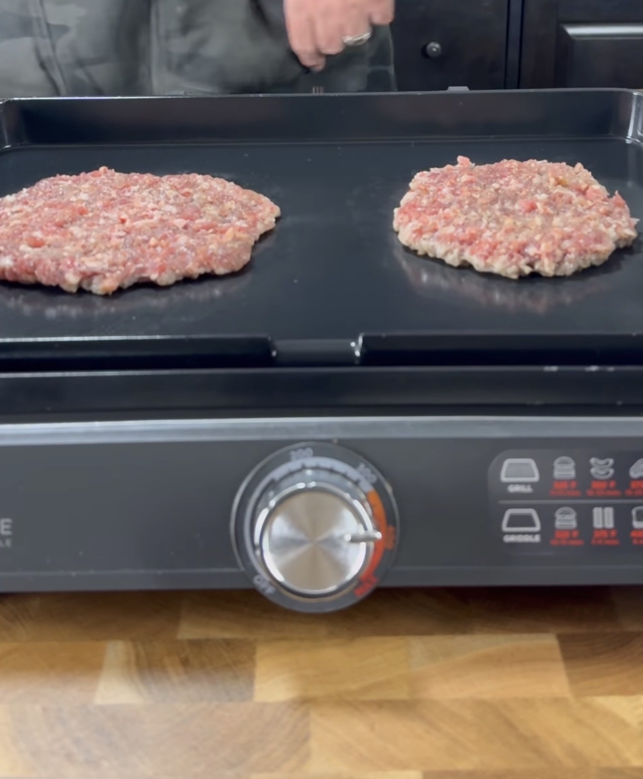 Two raw ground beef patties cooking on a black electric griddle with temperature dial and settings visible, set on a wooden countertop. A person stands in the background.