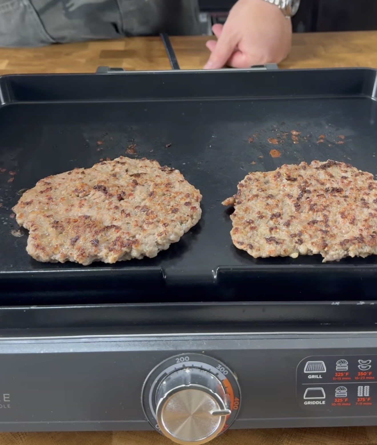Two hamburger patties are cooking on a black electric griddle. A persons hand is visible in the background above a wooden countertop. The griddle’s temperature control and settings are visible in the foreground.