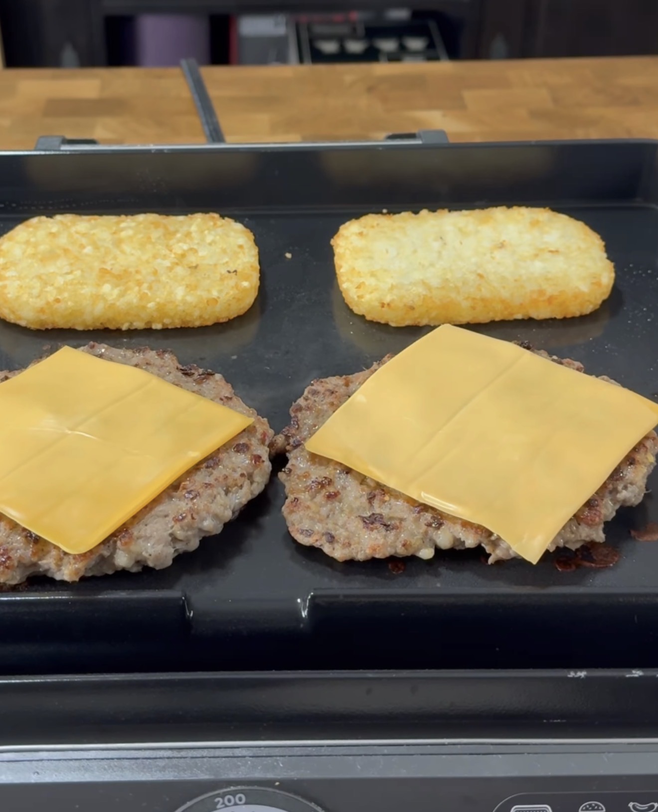 Two hash browns and two beef patties topped with cheddar cheese slices are cooking on a flat-top griddle. A wooden countertop and stove controls are visible in the background.