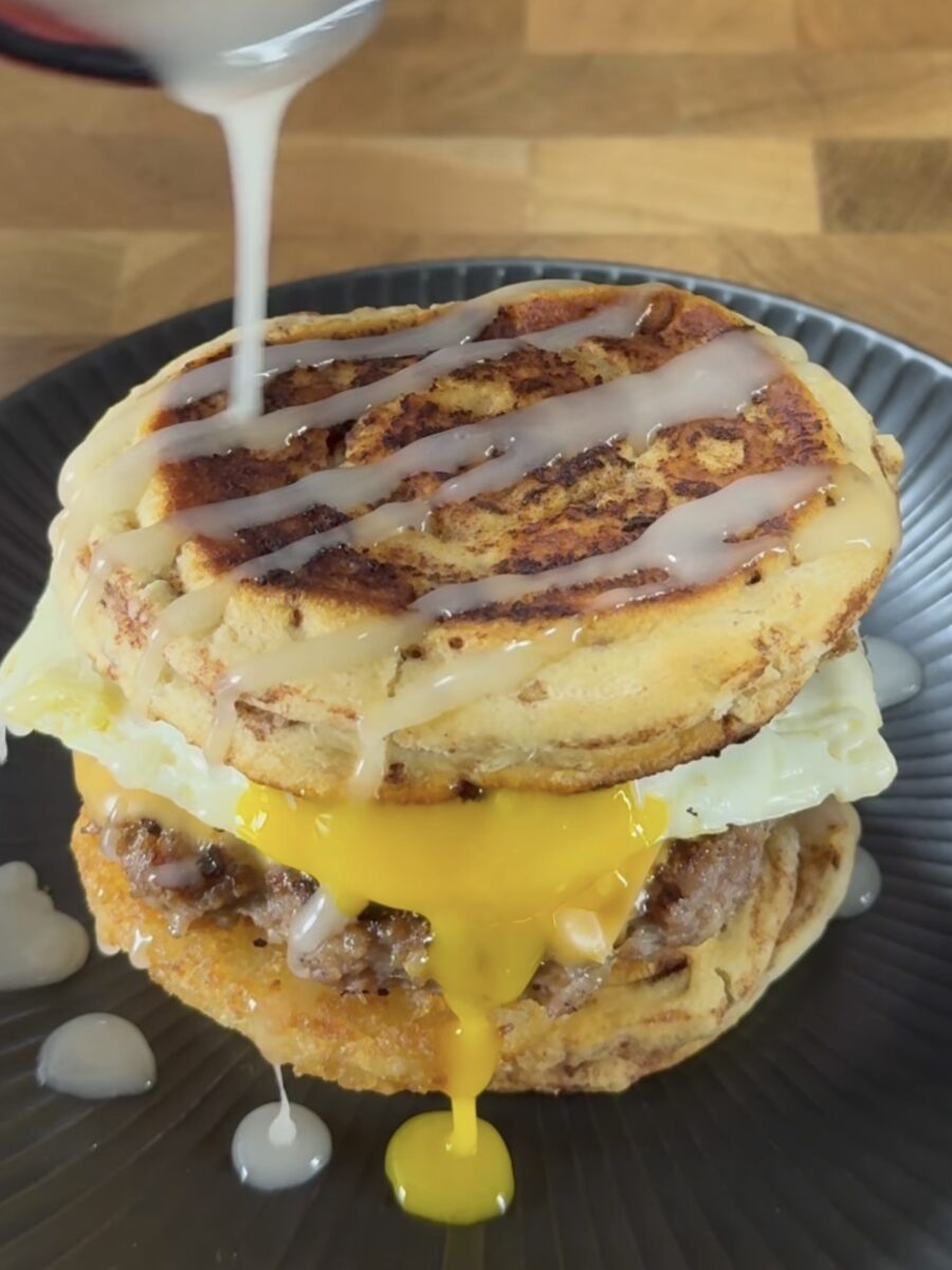 A breakfast sandwich with sausage, a fried egg with a runny yolk, and two thick, round pieces of French toast, being drizzled with syrup on a dark plate.