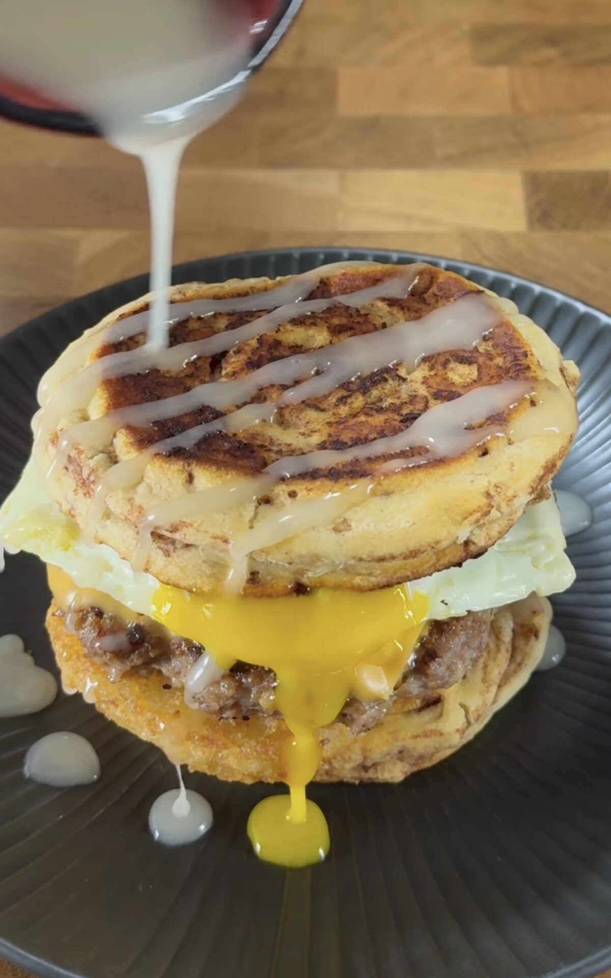 A breakfast sandwich with sausage, a fried egg, and two pancakes is topped with white icing being drizzled over it. The egg yolk is visibly running onto a dark plate beneath the sandwich.