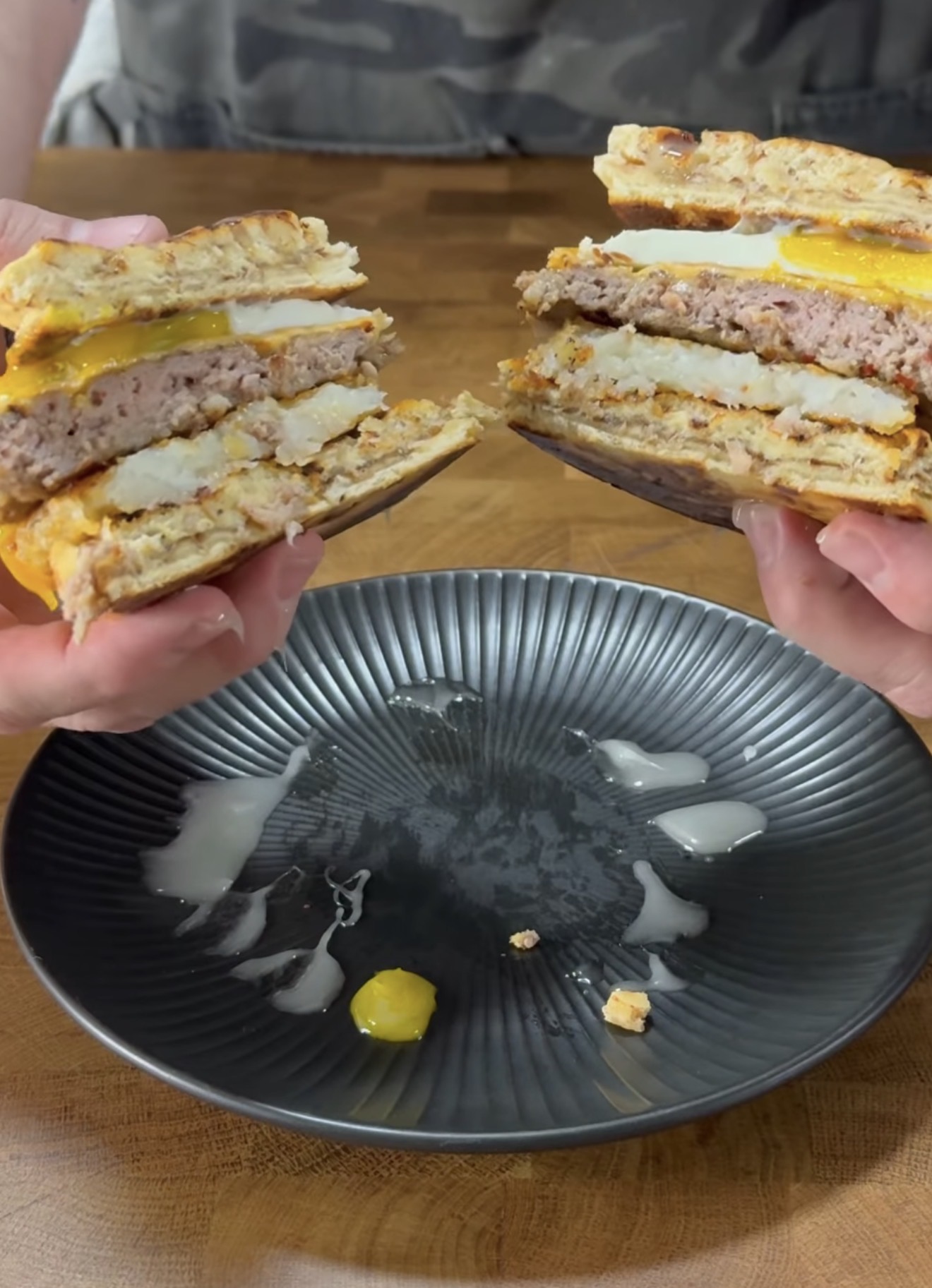 A person holds a breakfast sandwich split in half over a black plate. The sandwich has waffles, egg, cheese, sausage, and a hash brown, with some yolk and crumbs on the plate.