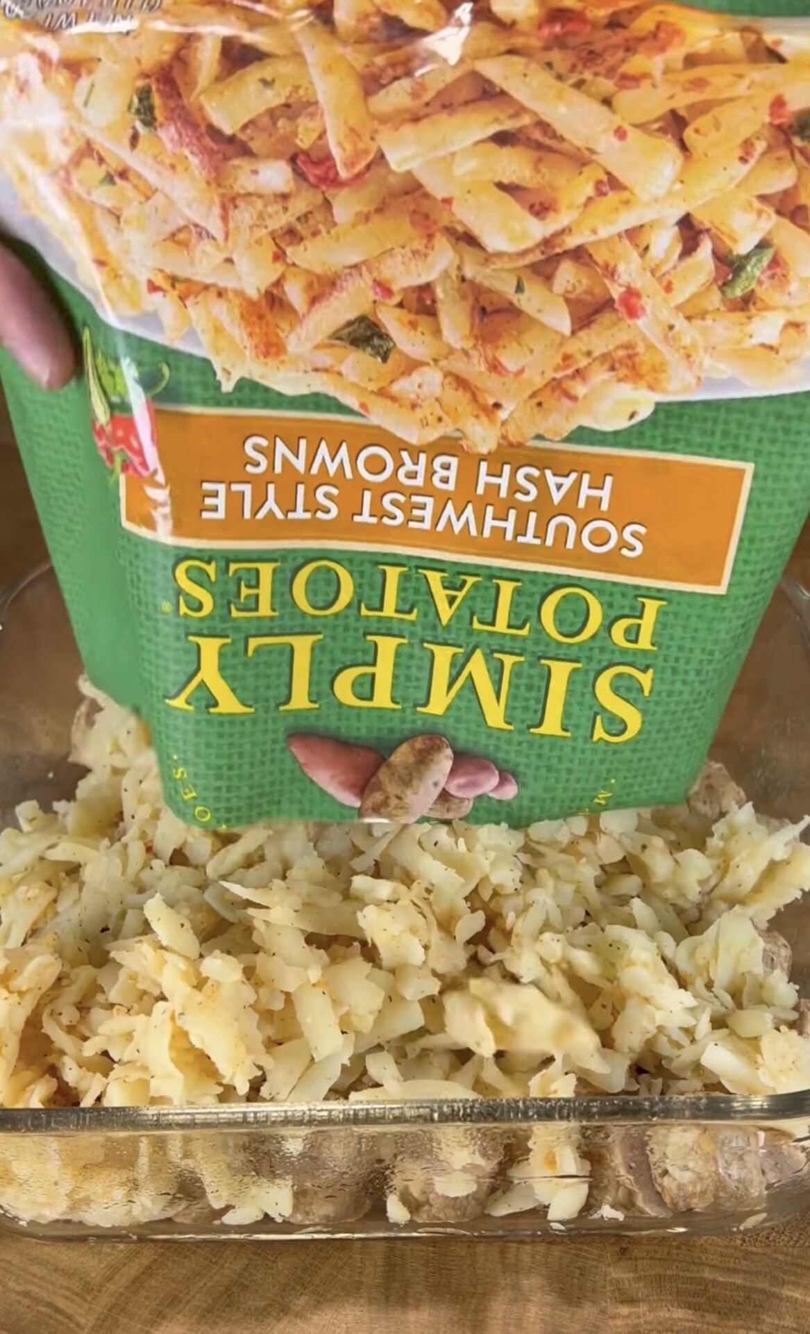 A hand pours frozen Southwest style hash browns from a green bag labeled Simply Potatoes into a glass baking dish.
