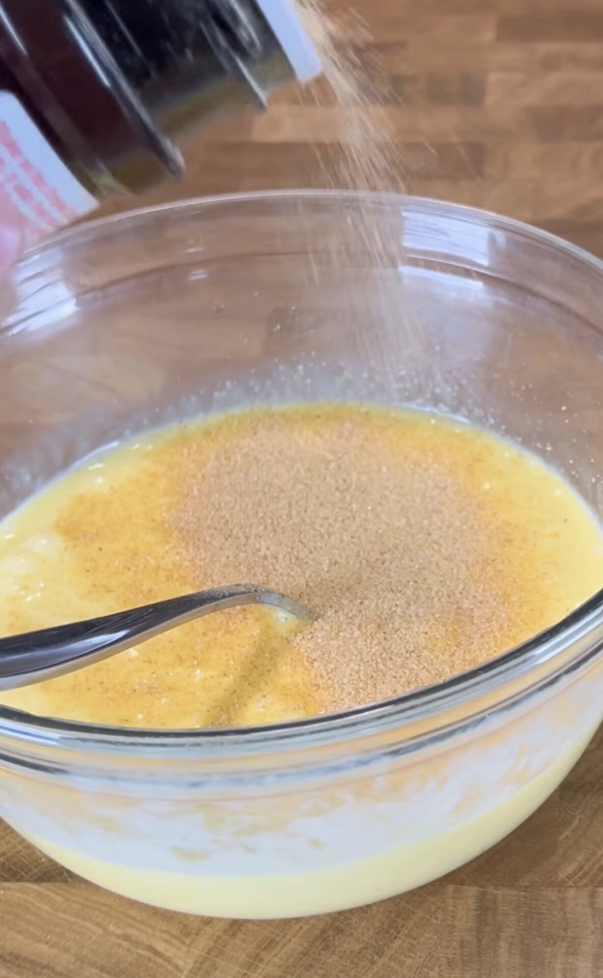 A spoon rests in a glass bowl filled with a yellow liquid mixture as seasoning is being sprinkled in from a container above. The bowl sits on a wooden surface.