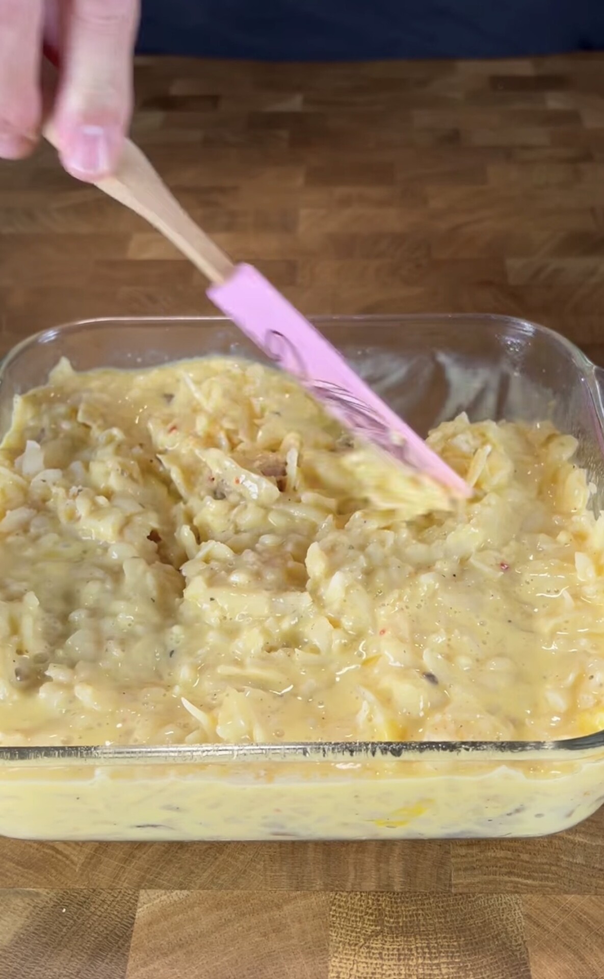 A hand uses a pink spatula to spread a creamy, cheesy potato mixture in a clear glass baking dish on a wooden surface.
