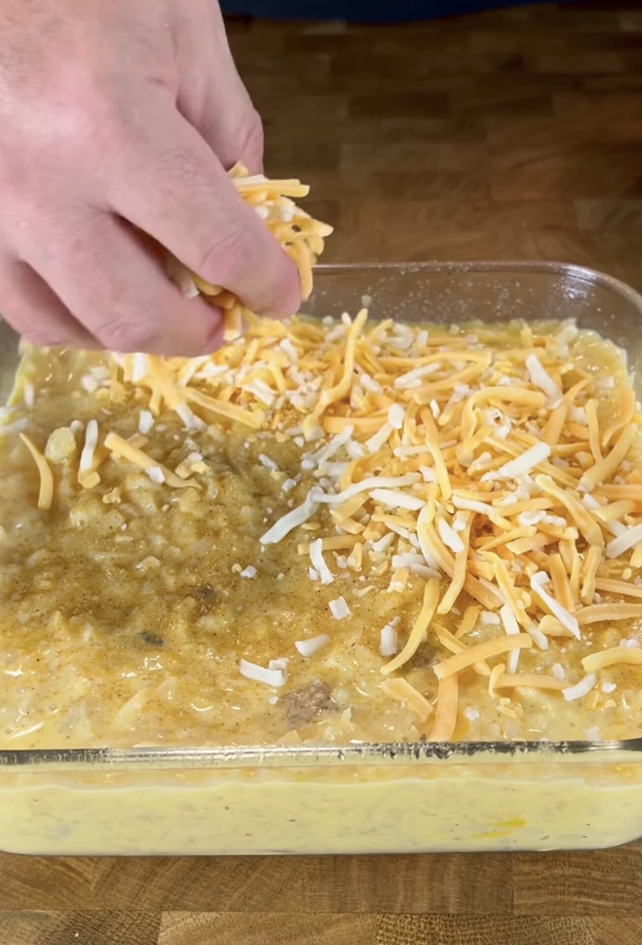 A hand sprinkles shredded cheese on top of a casserole mixture in a glass baking dish, set on a wooden surface.