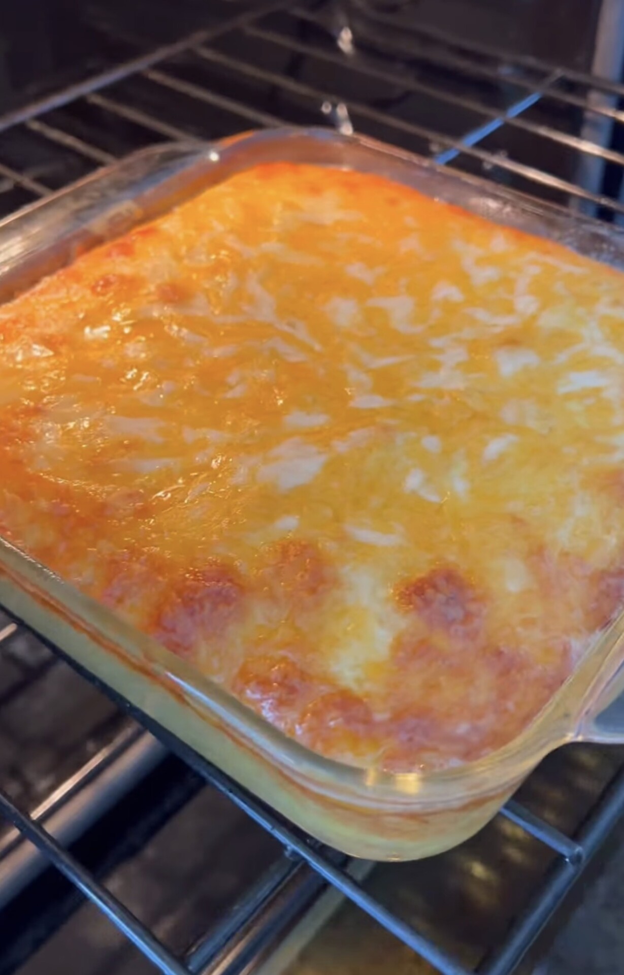 A glass baking dish filled with a golden, bubbly, and melted cheese casserole sits on an oven rack, with lightly browned spots across the surface.