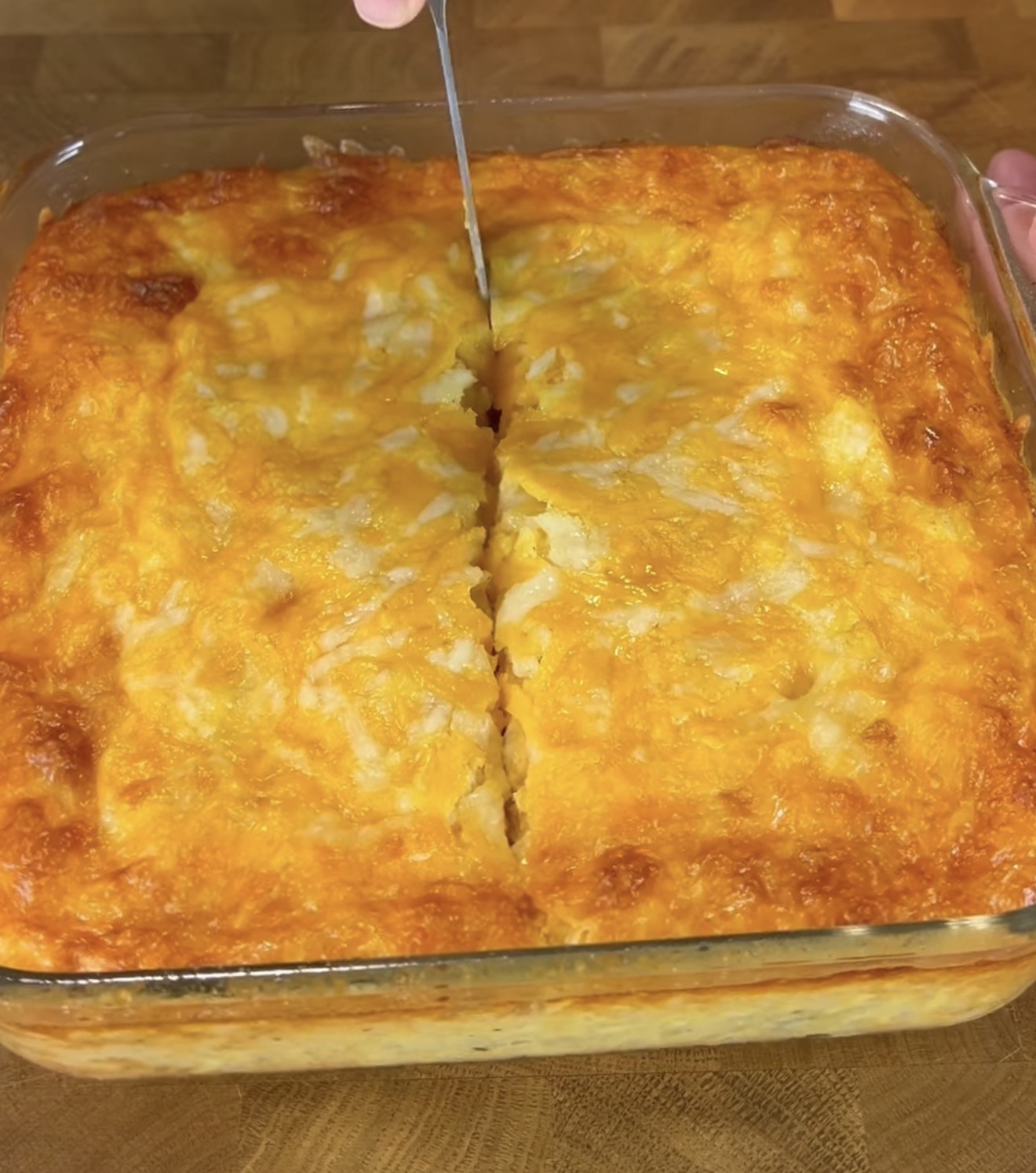 A hand slices into a freshly baked, golden-brown casserole topped with melted cheese in a glass baking dish on a wooden surface.