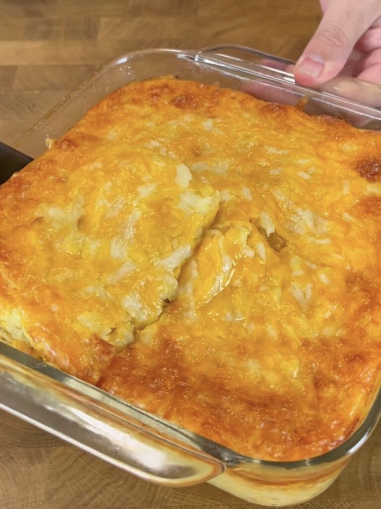 A hand holds a glass baking dish filled with a golden-brown, cheesy casserole, with a small section cut and ready to be served, resting on a wooden surface.