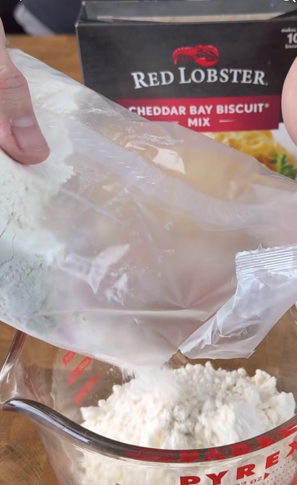 A hand pours Cheddar Bay Biscuit Mix from a clear plastic bag into a glass measuring cup, with a Red Lobster biscuit mix box in the background.