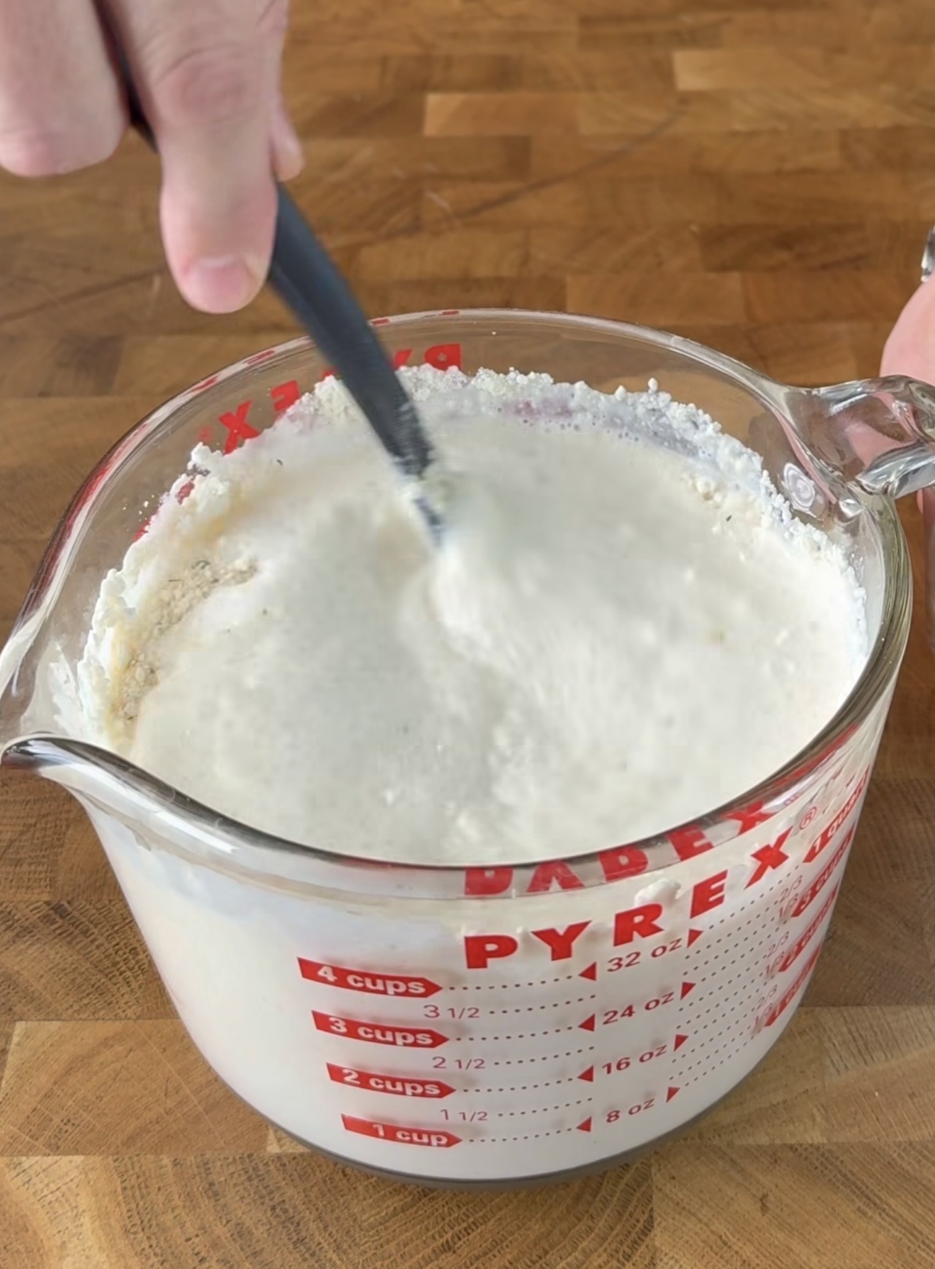 A hand stirs a thick, white mixture in a large glass Pyrex measuring cup with red markings, sitting on a wooden countertop.