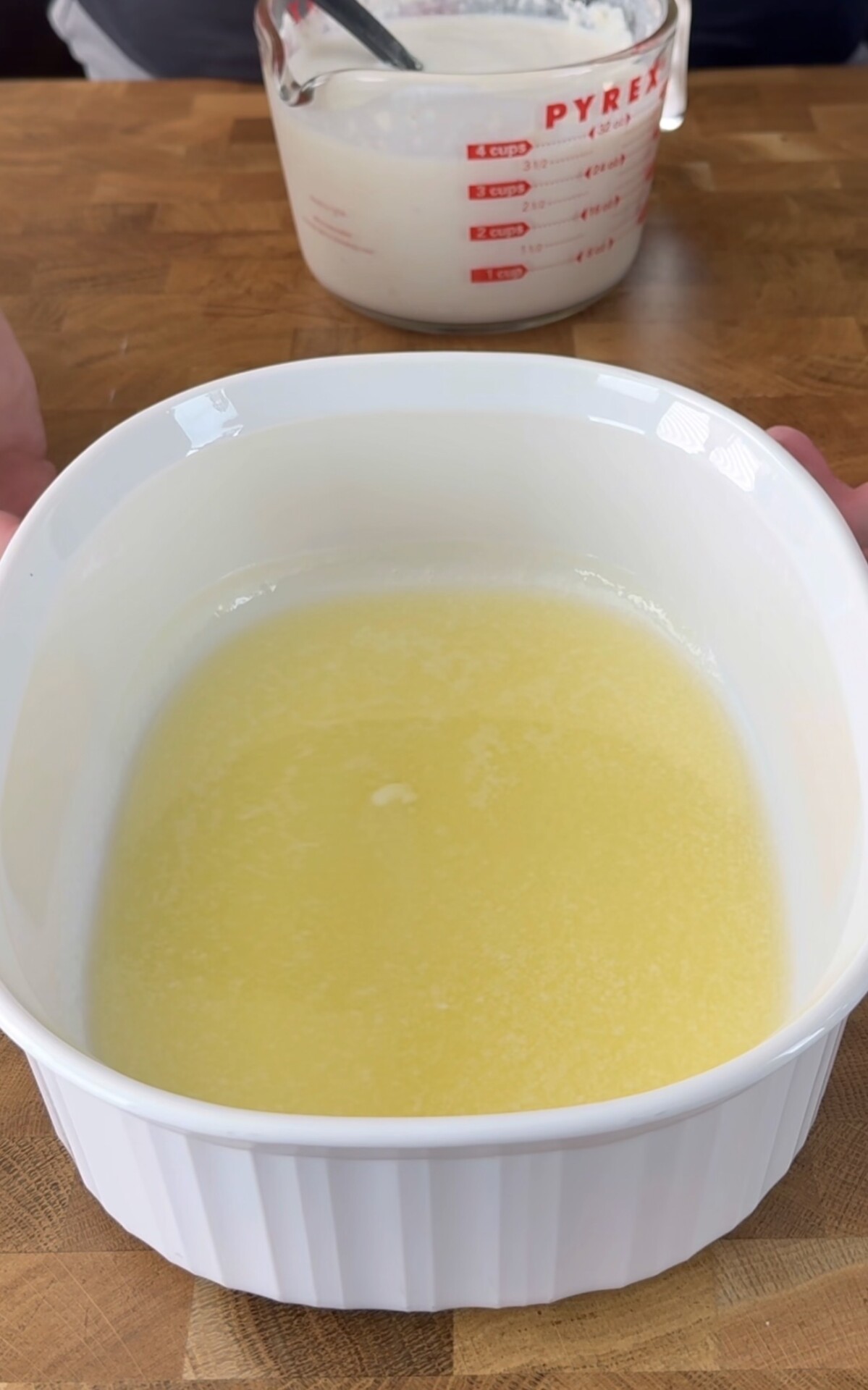 A white baking dish filled with melted butter sits on a wooden surface. In the background, a glass measuring cup with batter and a metal spoon is partially visible.
