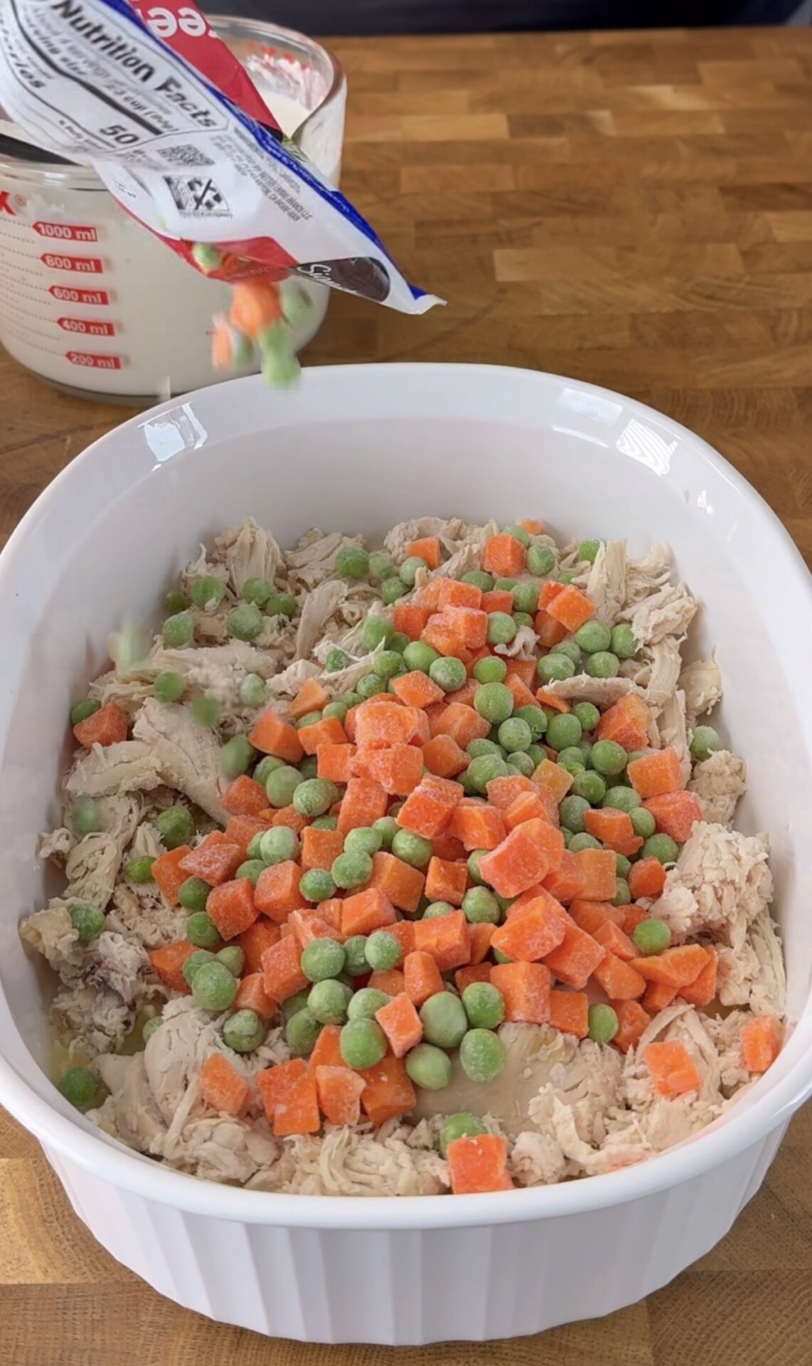 A white baking dish filled with shredded chicken is topped with frozen peas and diced carrots, being poured from a bag. A clear measuring cup with white liquid is in the background on a wooden countertop.