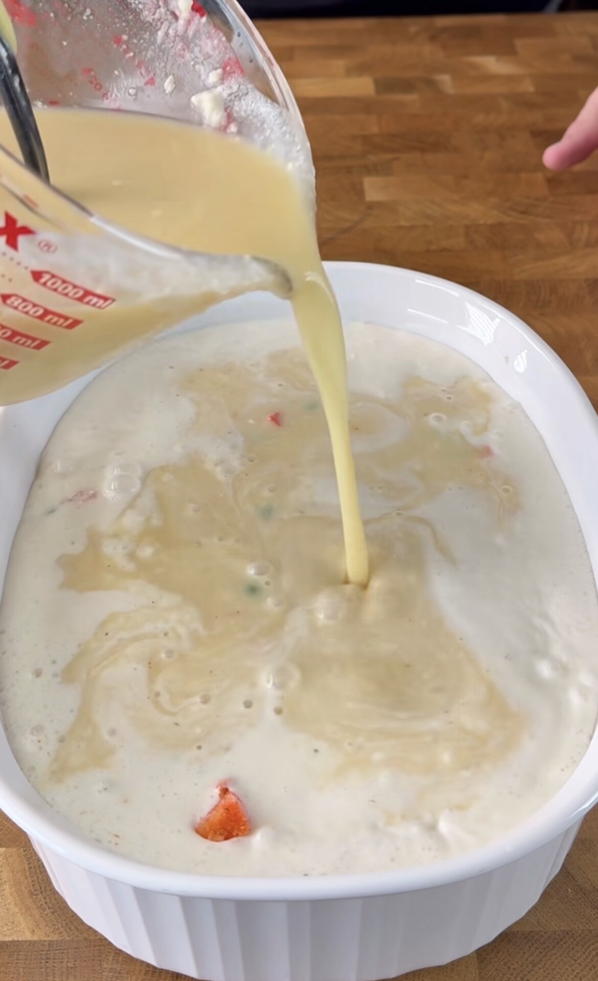A measuring cup pours a creamy yellow liquid mixture into a white casserole dish containing a partially mixed filling with visible vegetable pieces, on a wooden surface.