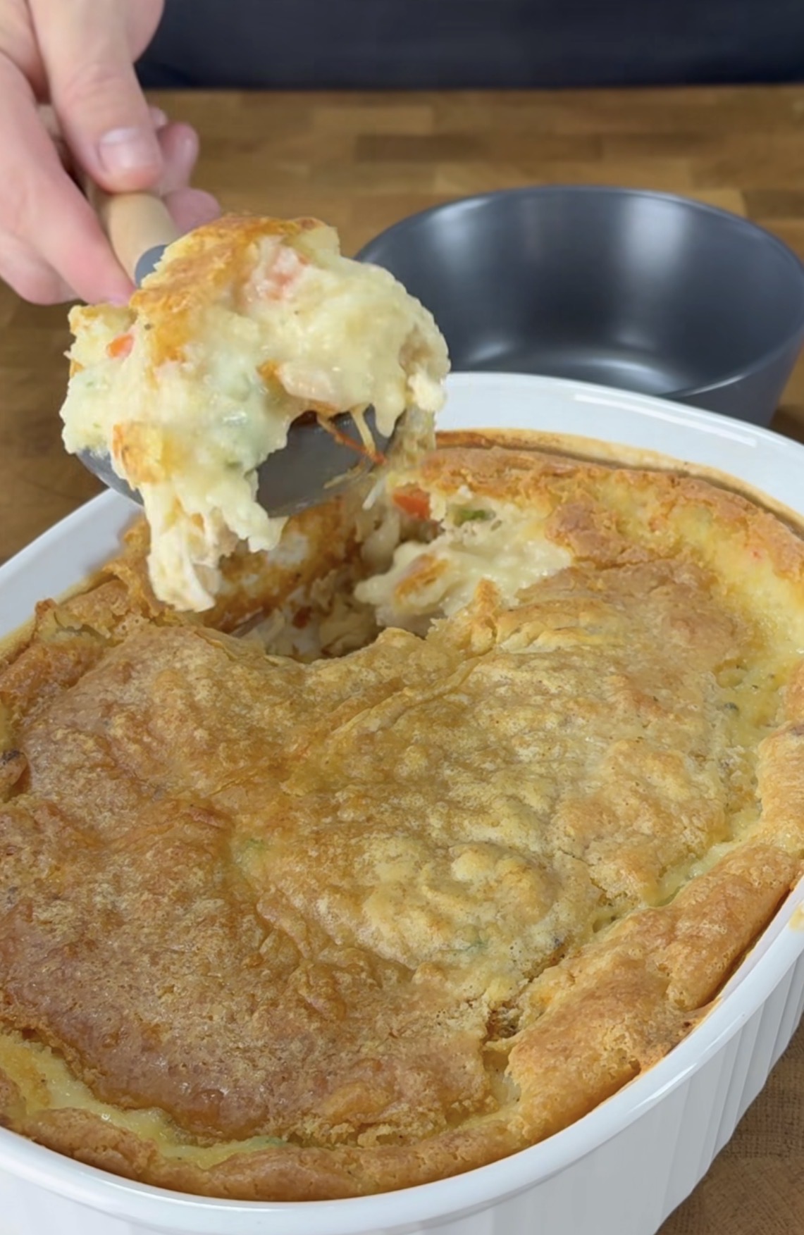 A hand serves a portion of creamy chicken pot pie with a golden, flaky crust from a white baking dish, with a dark gray bowl in the background.