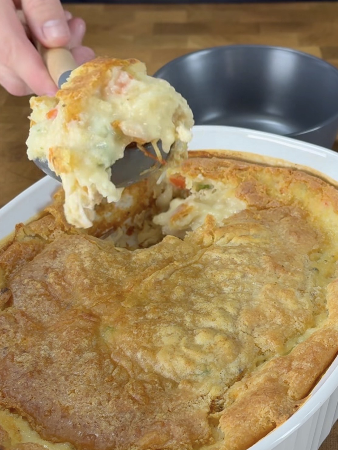 A hand scoops a serving of creamy chicken pot pie with a golden-brown crust from a white baking dish, with a metal bowl in the background.