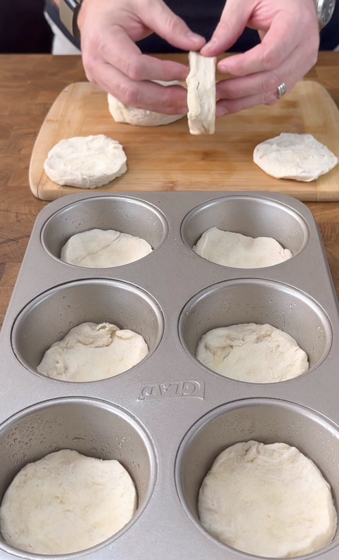 A person shapes biscuit dough rounds, placing them into a metal muffin tin, with more dough rounds on a wooden cutting board in the background.