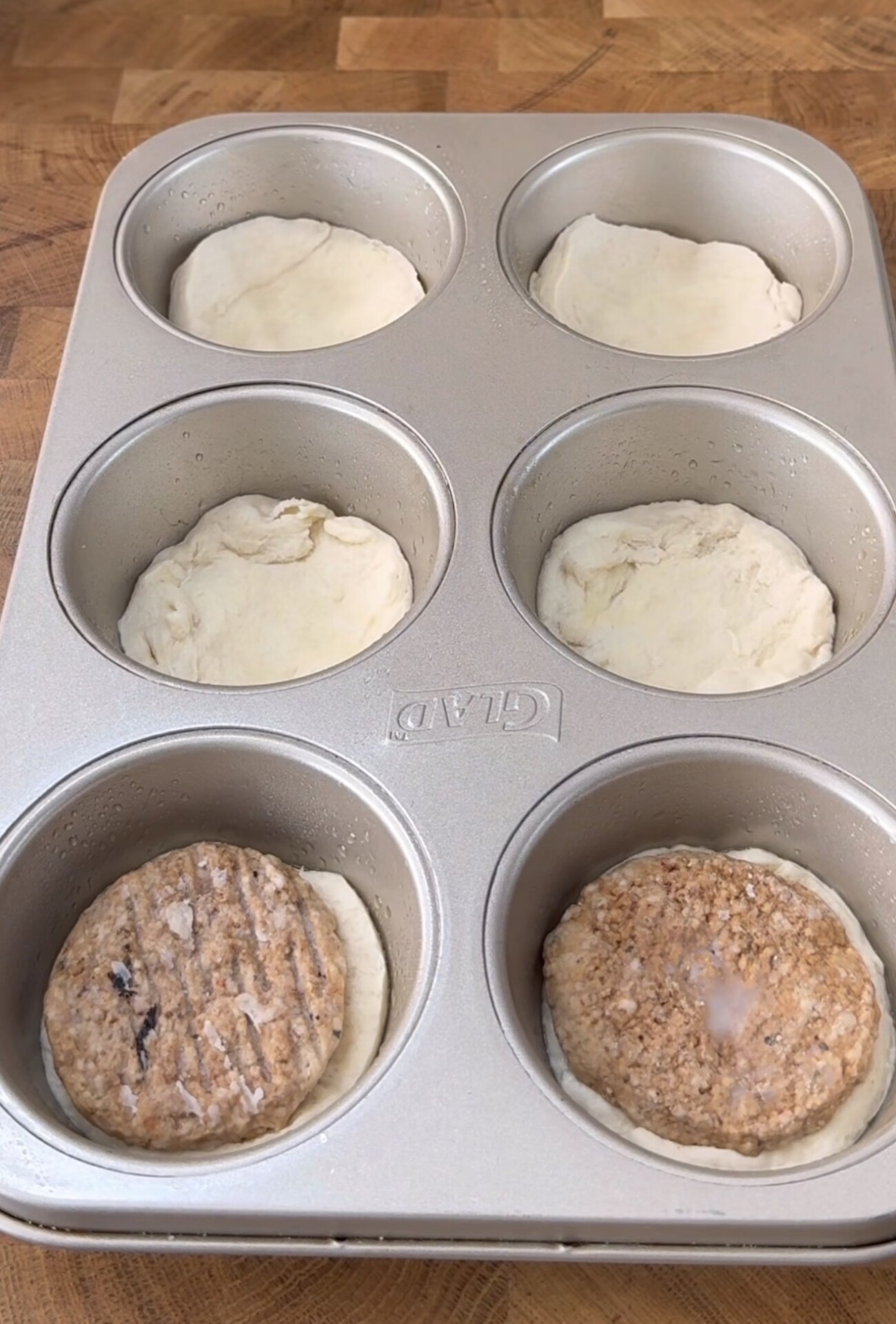 A muffin tin with six cups, each containing a round of dough. Two of the cups also have a frozen sausage patty placed on top of the dough. The tin is on a wooden surface.