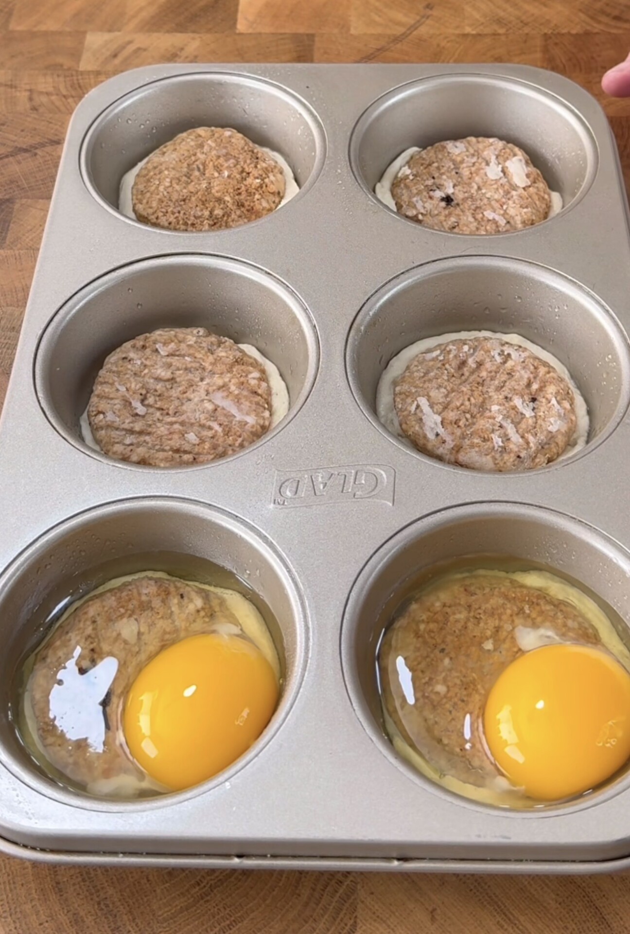 A muffin tin with six sections contains sausage patties in each, and three sections also have raw eggs cracked over the patties, all on a wooden surface.