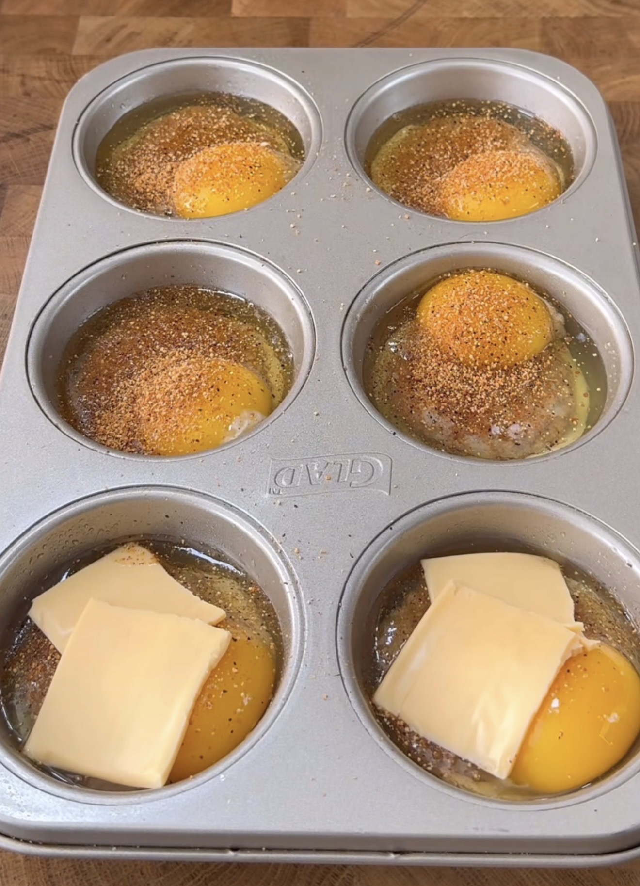 A muffin tin contains six sections, each holding a raw egg. Some eggs are topped with slices of yellow cheese, while others are sprinkled with seasoning. The tin is on a wooden surface.