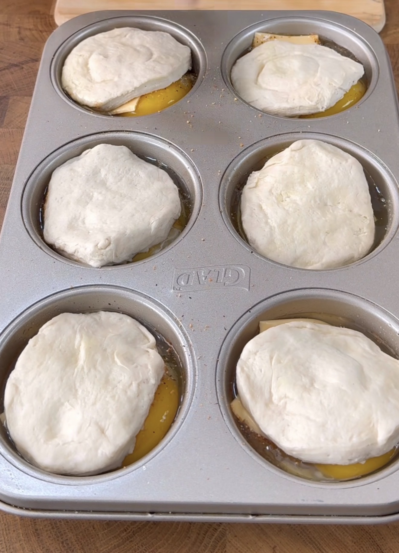 A muffin tin with six compartments, each filled with a layer of biscuit dough on top of melted butter and what appears to be cheese, ready to be baked.