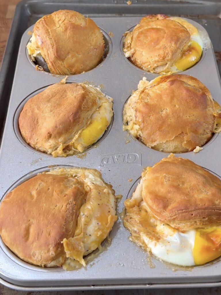 A muffin tin containing six baked biscuit cups, each filled with eggs and slightly oozing with filling. The tops are golden brown and some eggs are visible where the biscuit has separated.